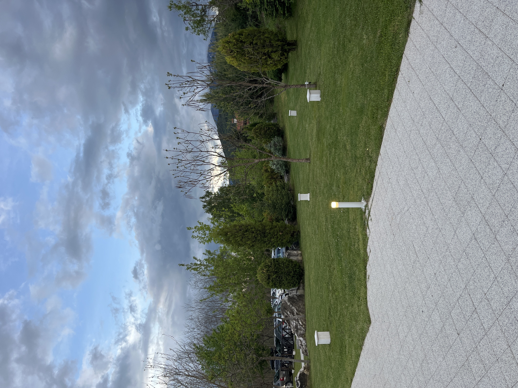 The image depicts a landscaped outdoor area in Koprivlen, Bulgaria, under a cloudy sky. A paved surface occupies a portion of the foreground, adjacent to a green lawn. The lawn is punctuated by several white, cuboid structures, likely lighting fixtures. Trees, some bare-branched, line the edge of the lawn, separating it from a parking area with multiple cars. Beyond the trees, a glimpse of mountains can be seen.