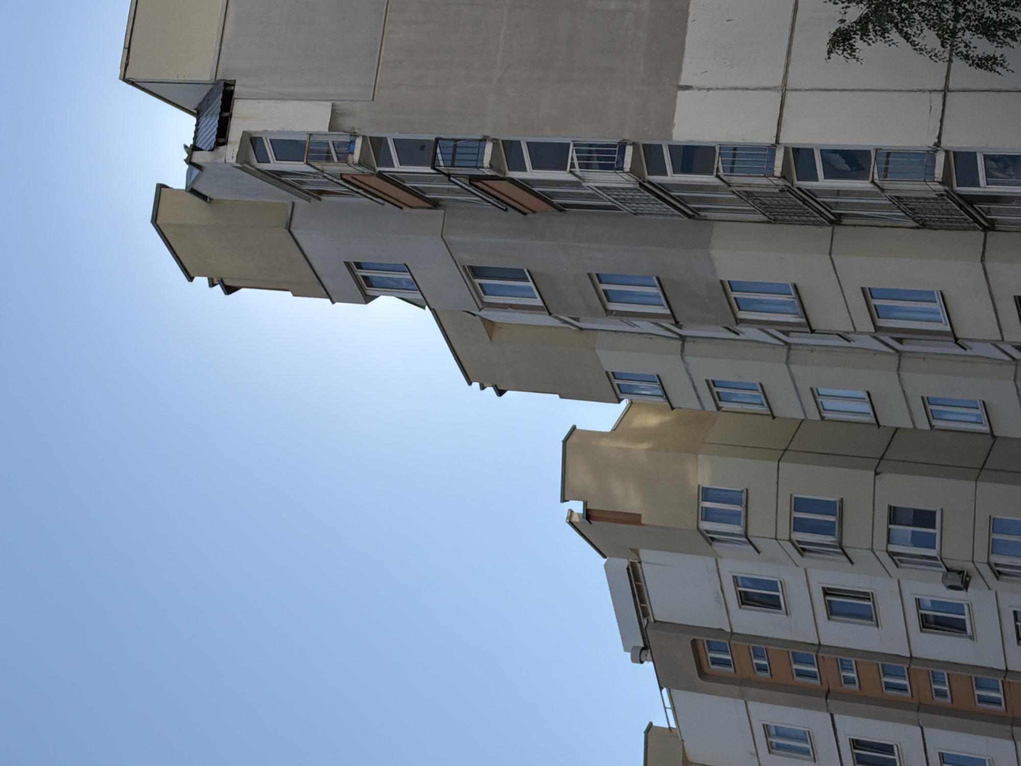 The image depicts a multi-story residential building under a clear blue sky. The building has a modern architectural style with a combination of beige and gray facades. Several windows and balconies are visible on the exterior. The perspective is from a low angle, looking up towards the top of the building. The lighting suggests it is daytime, with sunlight illuminating parts of the structure. The context provided indicates the location is Minsk, Belarus, suggesting a typical urban residential area in the city. There are no people or specific actions clearly depicted in the scene. The environment is dominated by the building and the sky.