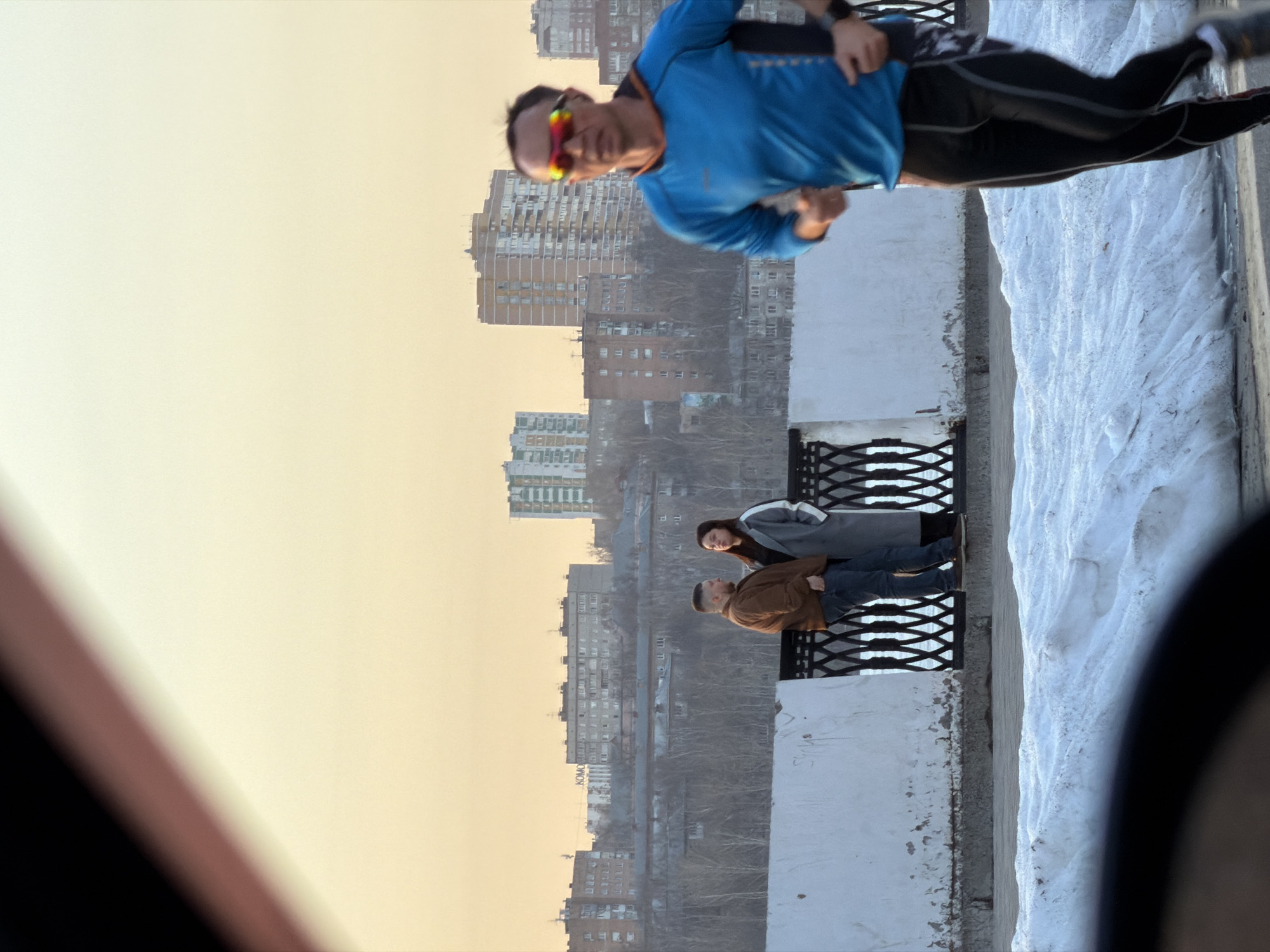 A horizontal outdoor shot depicts a cityscape, likely Izhevsk, Russia, under a pale yellow sky. In the foreground, a man in athletic attire – a bright blue long-sleeved shirt, black running tights, and sunglasses – is in motion, possibly jogging or running. He is positioned near a white-painted bridge or overpass with black metal railings. To his right, there's a stretch of snow-covered ground.
In the middle ground, two individuals are standing on the bridge or overpass. A man in a brown jacket is beside a woman in a grey coat. The backdrop reveals a dense urban environment with multiple apartment buildings and bare trees. In the lower right corner of the frame, part of an unidentified dark object is present, obscuring the view.