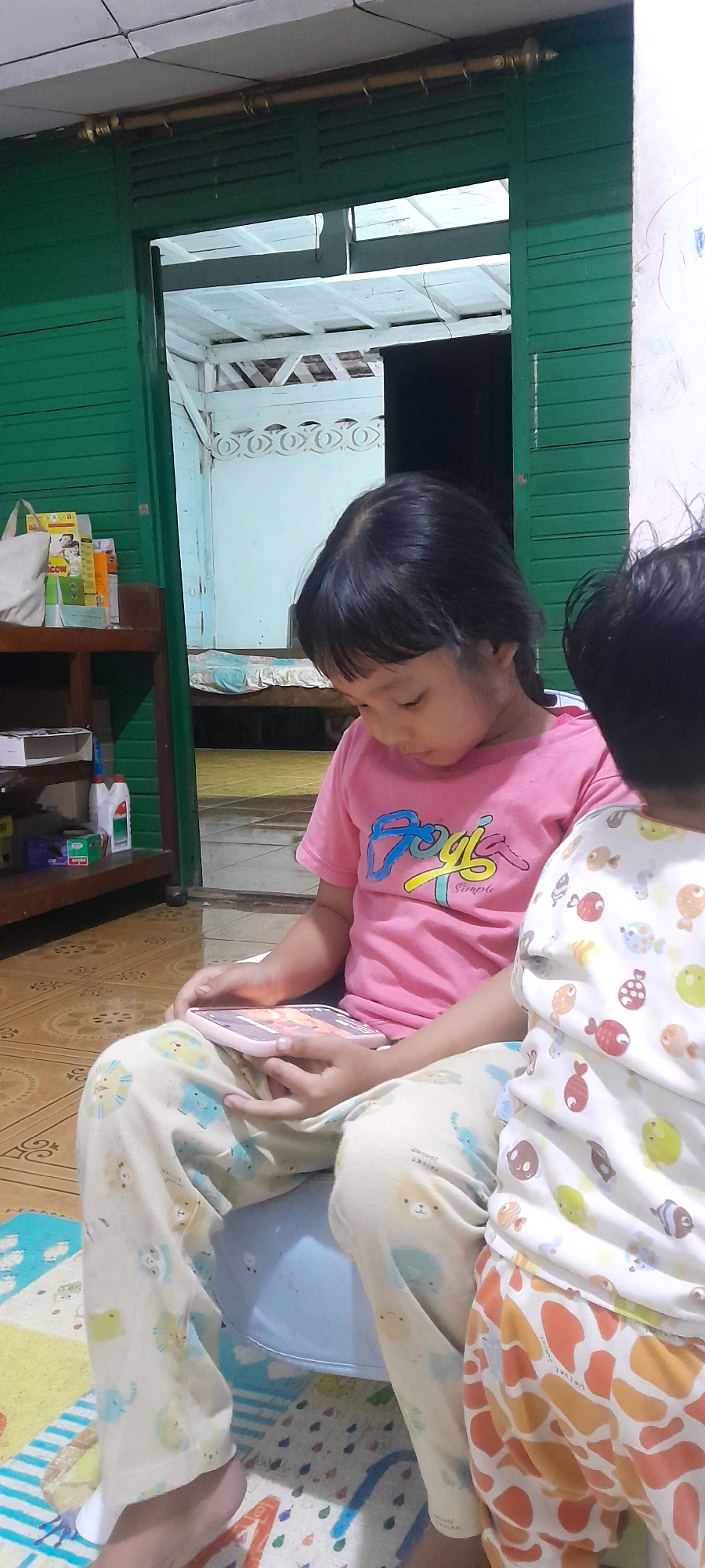 The image depicts an indoor scene, likely a residential interior, in Sambong, Indonesia. Two young children are seated, with one child in the foreground looking down at a tablet device held in their lap. This child is wearing a pink t-shirt with colorful lettering and light-colored pants adorned with cartoon animal patterns. Next to them, another child, partially visible, wears a white shirt with a fish pattern and orange and white patterned pants. They are positioned on a floor covered with a patterned mat. In the background, a doorway with green wooden doors opens into another room with a white ceiling and wooden beams. A shelf with various items, including bottles and boxes, is visible to the left. The overall setting appears casual and domestic.