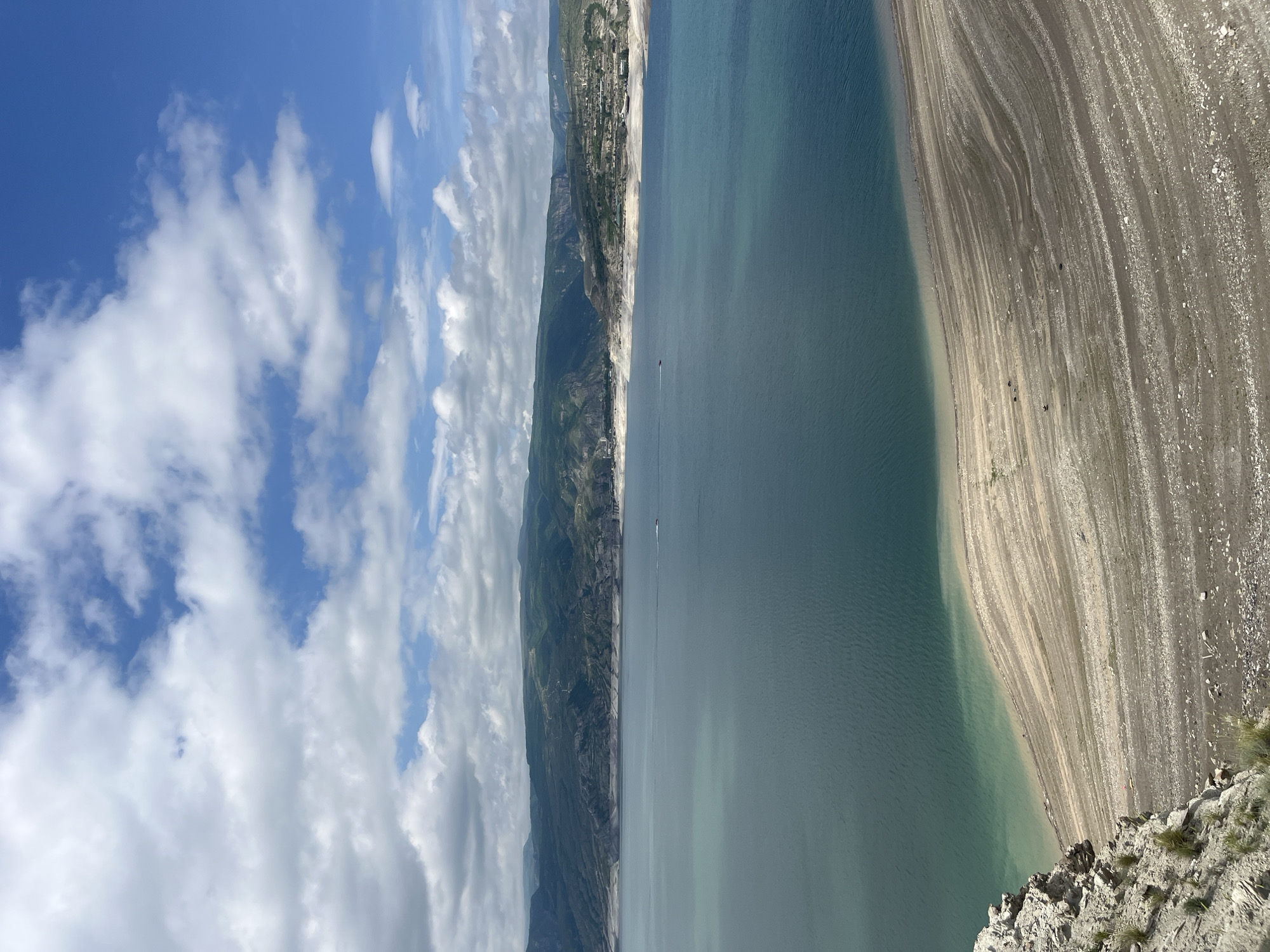 The image shows a landscape with a body of water, potentially a lake or reservoir, alongside a broad, exposed shoreline that appears to be rocky and sandy. The opposite side of the water features cliffs and hills, with some development at the top of the cliffs. The sky is partially cloudy. The scene could be near Chirkey, Russia, given the likely landscape context. The image does not have people present.
