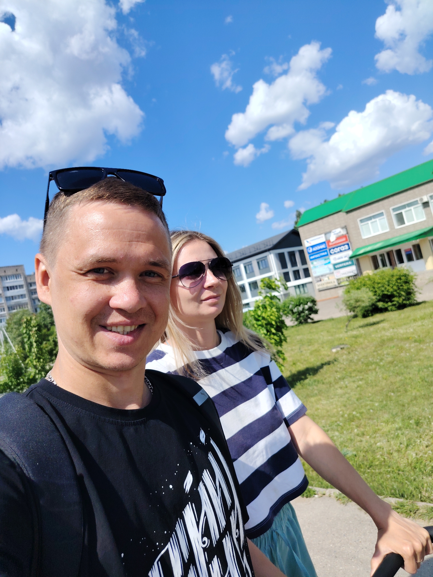 A selfie depicts a man and a woman, likely a couple, outdoors on a sunny day. The man is in the foreground, smiling at the camera, wearing a black t-shirt and sunglasses on his head. The woman is behind him, also wearing sunglasses and a blue and white striped shirt. They appear to be on a paved path near a grassy area.
In the background is a building with a green roof and signage in Russian. One sign contains the word "Союз," which transliterates to "Soyuz," and another sign indicates "Електрика." The building suggests a commercial or office space. There is another modern-looking building visible in the background. The sky is blue with scattered white clouds. Based on the signage and architectural style, the location is Chaikovsky, Russia.