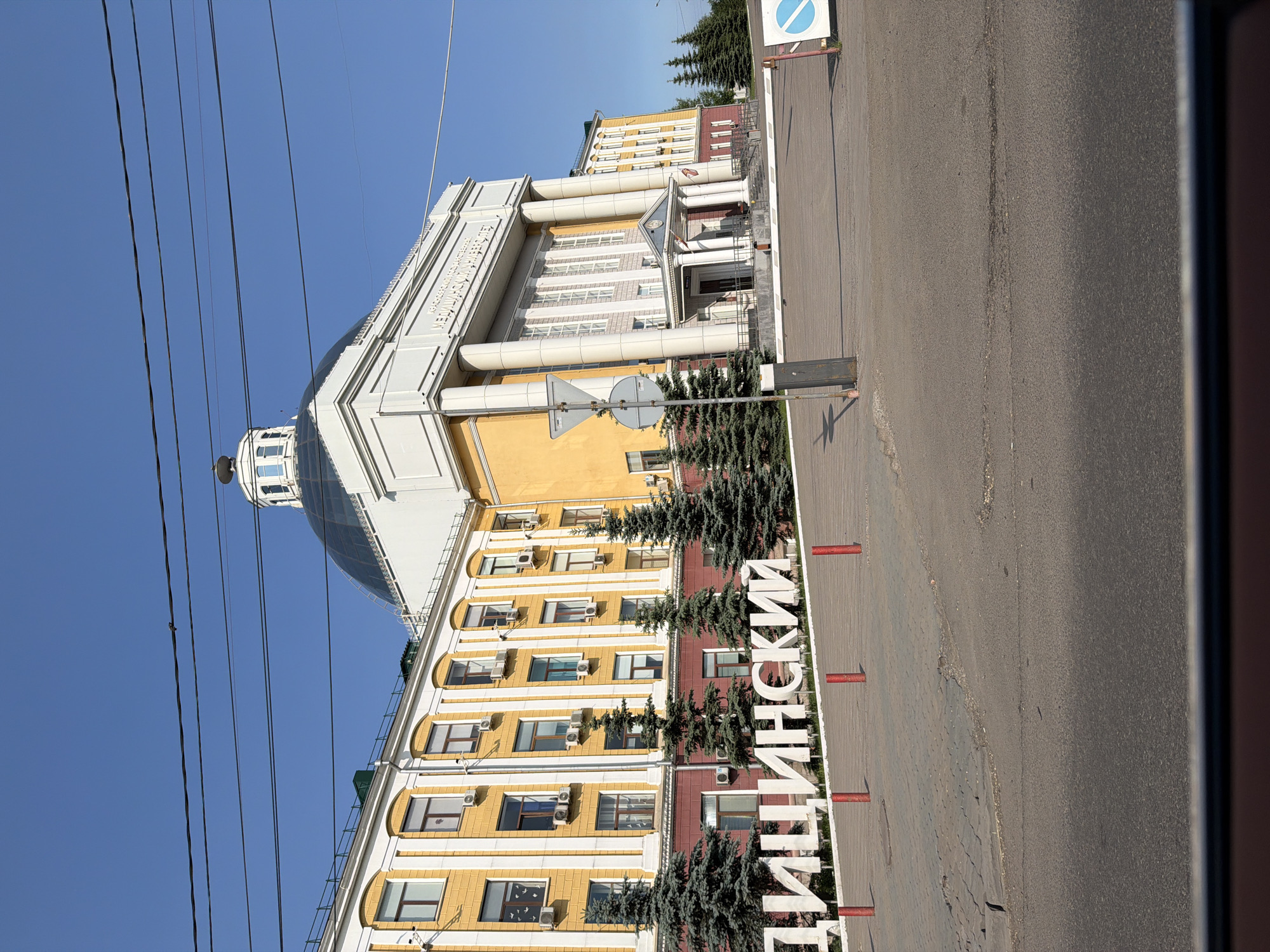 The image shows the Krasnoyarsk State Medical University named after Professor V.F. Voino-Yasenetsky in Krasnoyarsk, Russia. The building is large, with a yellow facade and white trim. It has a prominent dome and is surrounded by fir trees. In the foreground, large white letters spell out "МЕДИЦИНСКИЙ" (Medical). A road and sidewalk are visible, along with a barrier and some traffic signs. The sky is clear and blue.
