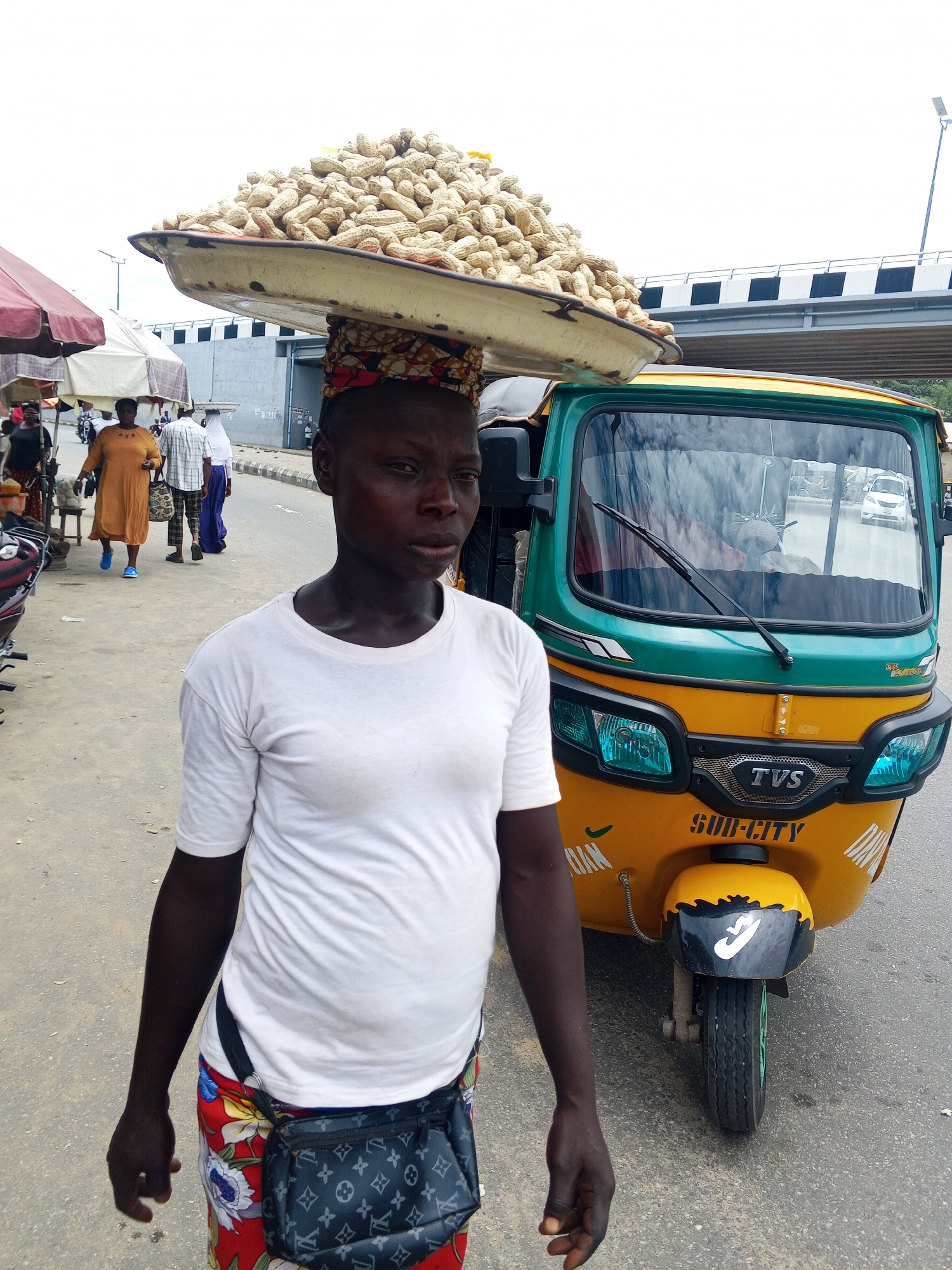 An individual stands on a paved road in Lokoja, Nigeria, carrying a large tray of peanuts balanced on their head. They wear a white t-shirt and floral-patterned pants, with a black fanny pack around their waist. To the right of the individual is a yellow and green auto-rickshaw with the logo "TVS" on the front and "SUB-CITY" written below it. In the background, a pedestrian walkway is visible, along with a bridge and a few other people and a motorcycle. The sky is overcast.