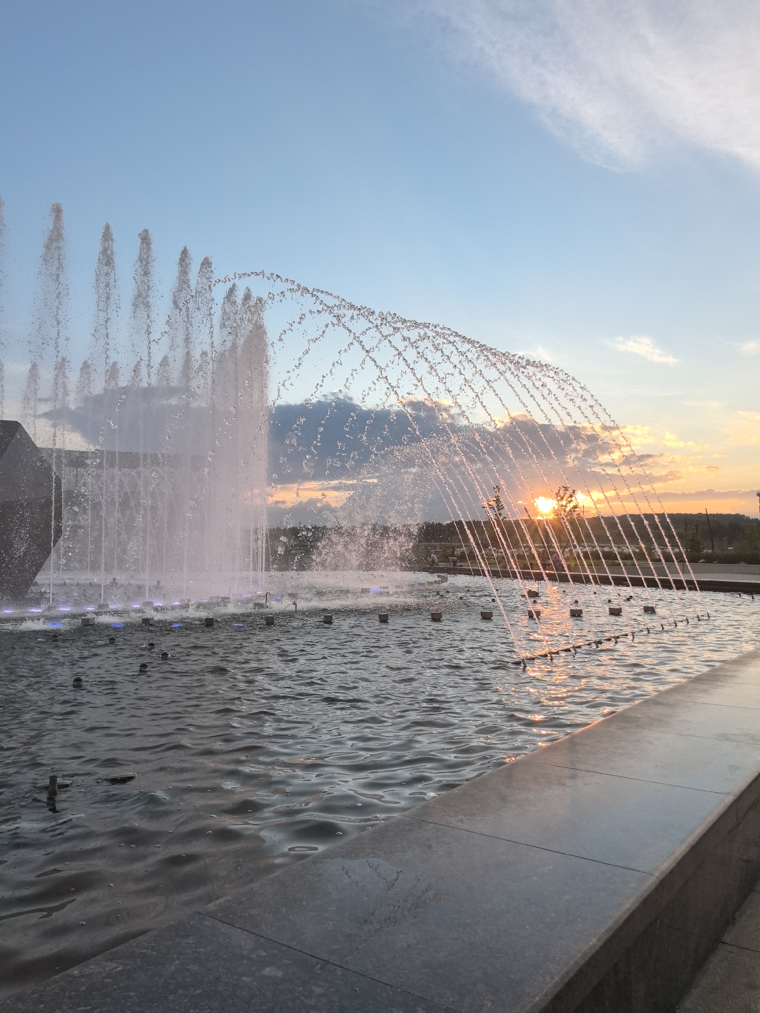 The image depicts a sunset over a large fountain. The fountain is in the foreground, with streams of water arching upwards and catching the golden light of the setting sun. The water is rippling, and several small nozzles are visible just below the surface. In the background, there is a line of trees and a hill, with a few clouds in the sky. The sky transitions from a clear blue at the top to a warm orange and pink near the horizon. The image appears to have been taken from a slightly elevated position, looking across the fountain.
The location is the Fountain "Youth" in Magnitogorsk, Russia.