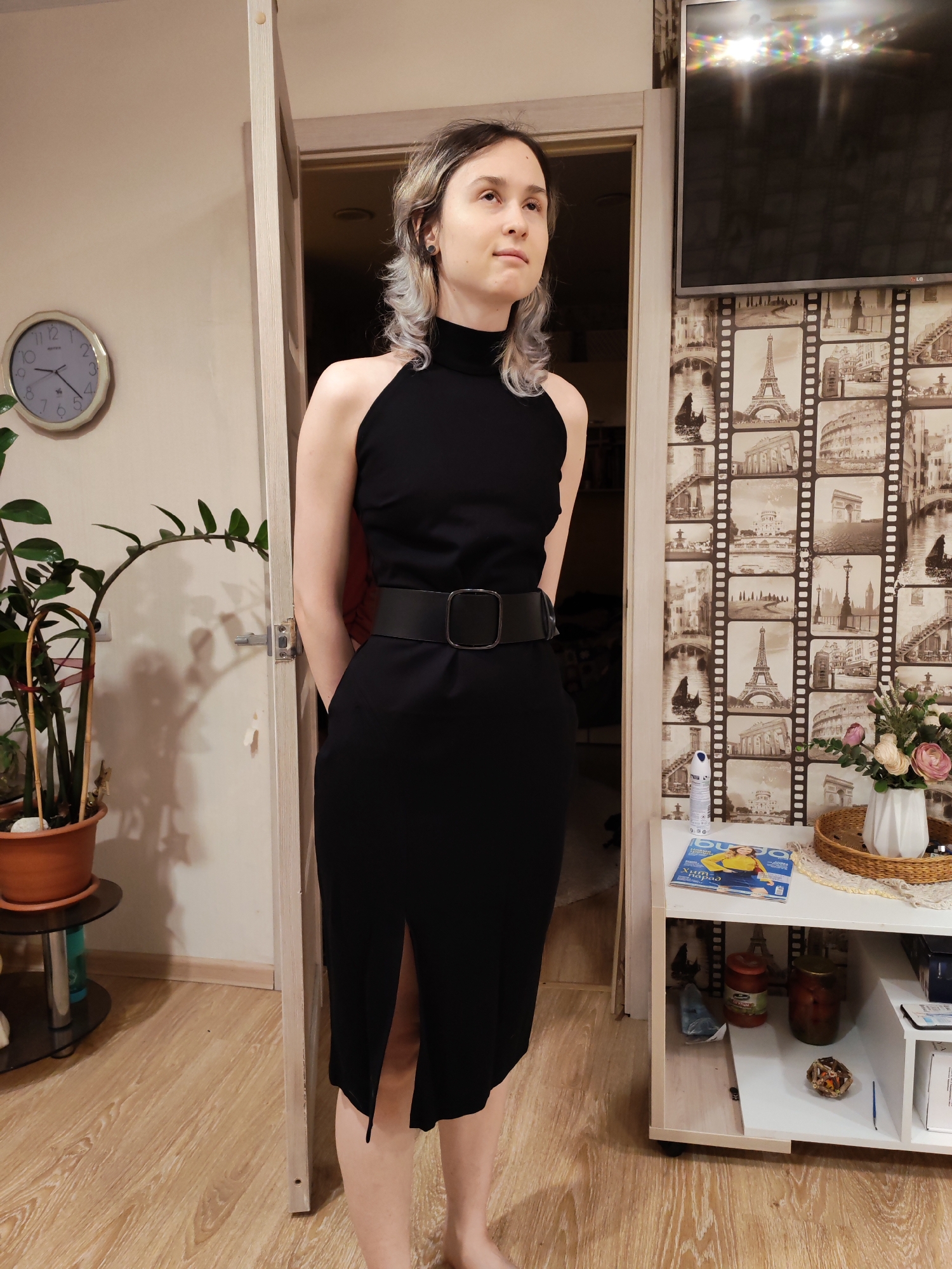 A young woman stands in a residential interior, likely in Chaikovsky, Russia, in a full-body shot. She has light-colored, wavy hair and wears a sleeveless, high-neck black dress with a thick black belt, her hands behind her back. Her gaze is directed slightly upwards and to her right, with a neutral, contemplative expression.
The room features light-colored walls and a light wood-laminate floor. To the woman's left, a round wall clock with "MAESTRO" visible on its face shows approximately 4:45. Beneath the clock, a tall green Zamioculcas zamiifolia (ZZ plant) in a brown pot rests on a black and glass stand.
Behind and to the right of the woman, a wall is adorned with decorative wallpaper featuring a sepia-toned filmstrip pattern of various European landmarks, including the Eiffel Tower, the Colosseum, and the Arc de Triomphe. Above this wallpaper, an LG flat-screen television is mounted, reflecting bright overhead lights.
To the woman's right, a white shelving unit holds several items: a woven tray with a white vase containing artificial pink and white flowers, an air freshener spray can, and a magazine with Cyrillic text that reads "Журнал 'ХИТ-ПАРАД'". On a lower shelf of the unit, two jars of preserves or pickles are visible. The artificial lighting and reflections suggest the photo was taken indoors during the evening or night. The overall setting is a modern, decorated home.