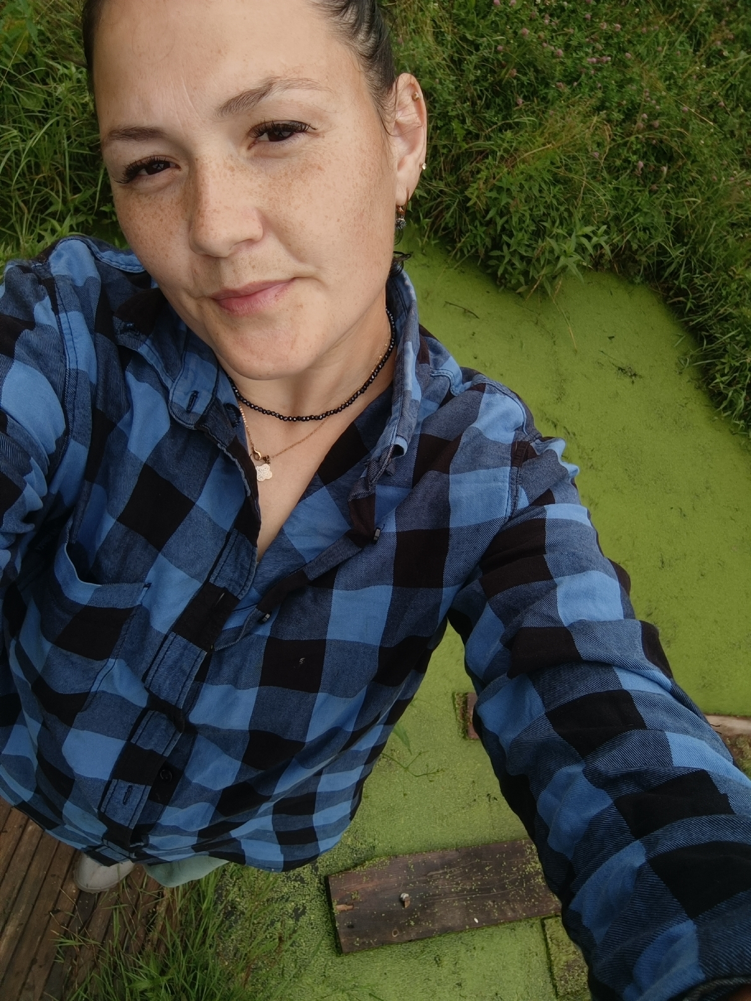 A woman with freckles and dark hair tied back is taking a selfie. She is wearing a blue and black plaid long-sleeved shirt and a black beaded necklace with a small gold pendant. She appears to be standing on a wooden surface, possibly a deck or pier, next to a body of green, algae-covered water. Tall green grass and some small pink flowers are visible in the background. The image is a close-up, focusing on the woman from the chest up. There are no discernible landmarks to indicate the specific location within the city of Karsha, Russia.