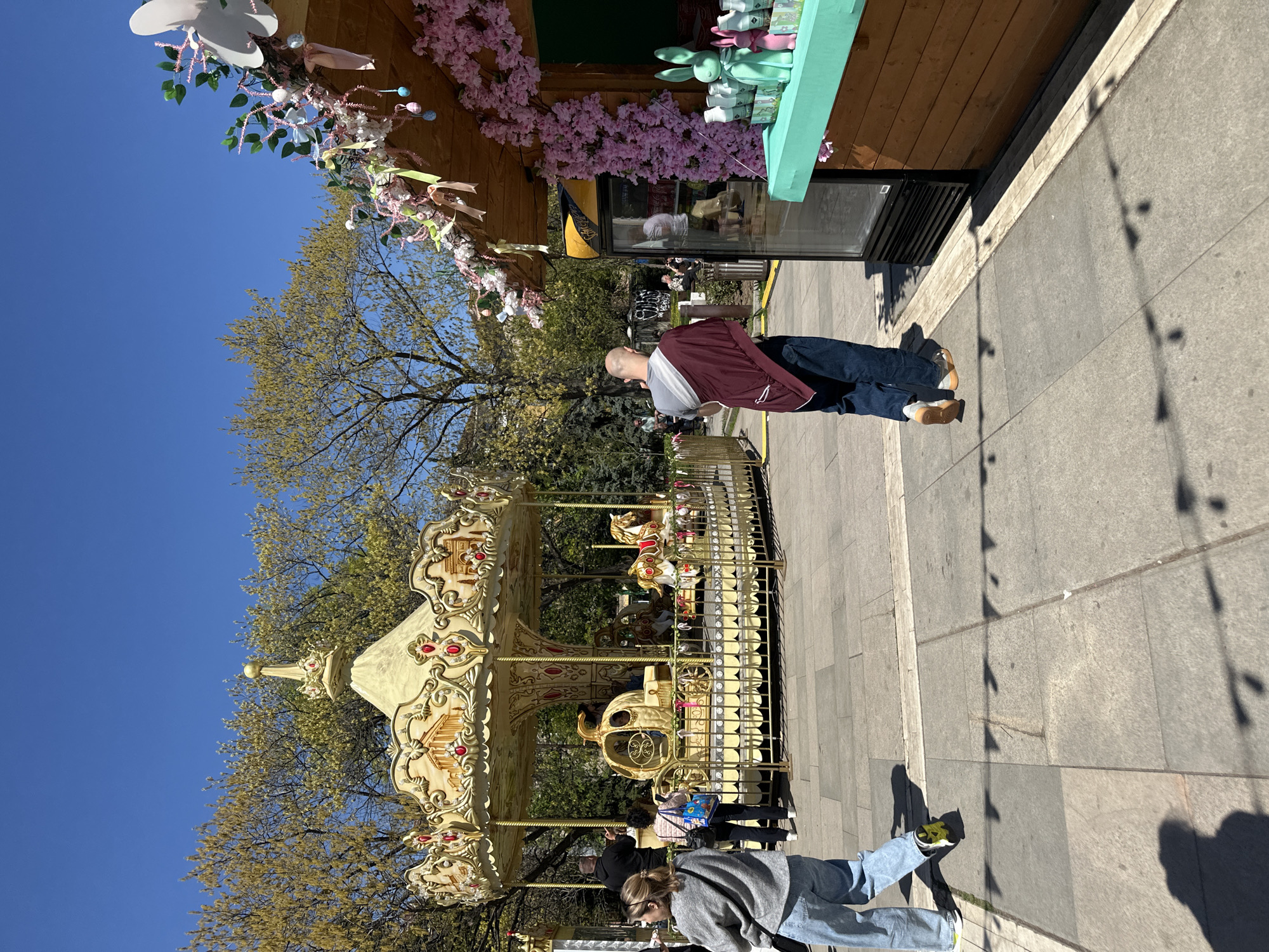 The image depicts a carousel in Sofia, Bulgaria, with a person standing nearby. The carousel is ornate and gilded, with a backdrop of trees and a clear blue sky. A person is standing close to the carousel. A wooden structure decorated with flowers and toy bunnies is visible, probably a sales kiosk. The ground is paved with gray stones. The image appears to be taken during the daytime, with strong shadows indicating direct sunlight.