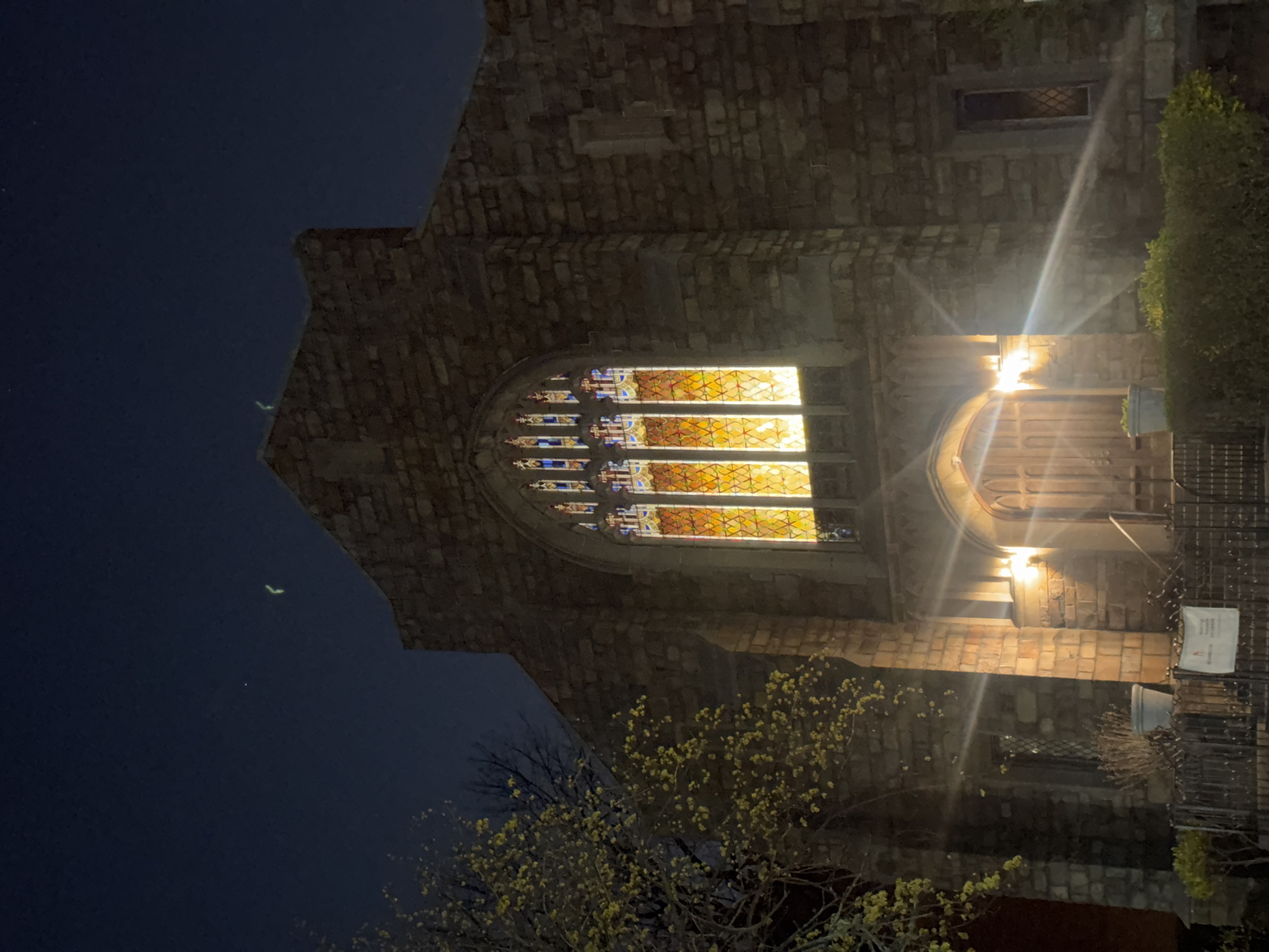 The image captures the exterior of a stone building, likely a church, at night. A large arched window with stained glass emits a warm, golden light. The walls are constructed of rough-hewn stones. Trees are visible in the foreground, some with small yellow flowers. A sign is attached to a fence on the right side of the frame. Given the architectural style and context, this could be a church in New York City, United States of America.