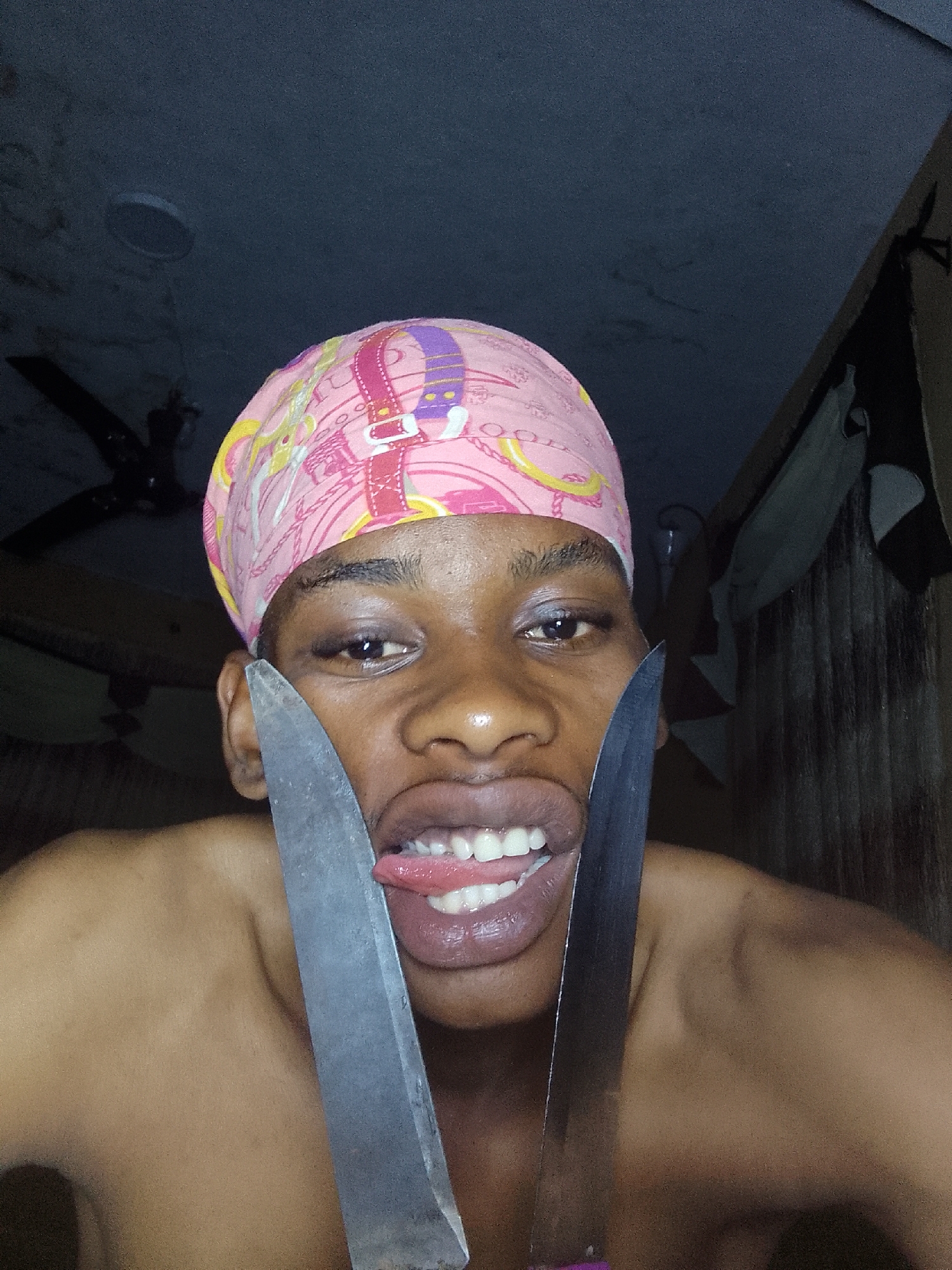 A person with dark skin and a pink patterned headscarf is holding two knives up to their face, with their tongue sticking out between the blades. The person is shirtless. The background appears to be a dark room with a ceiling fan and some draped fabric. The location is Unknown, Unknown.
