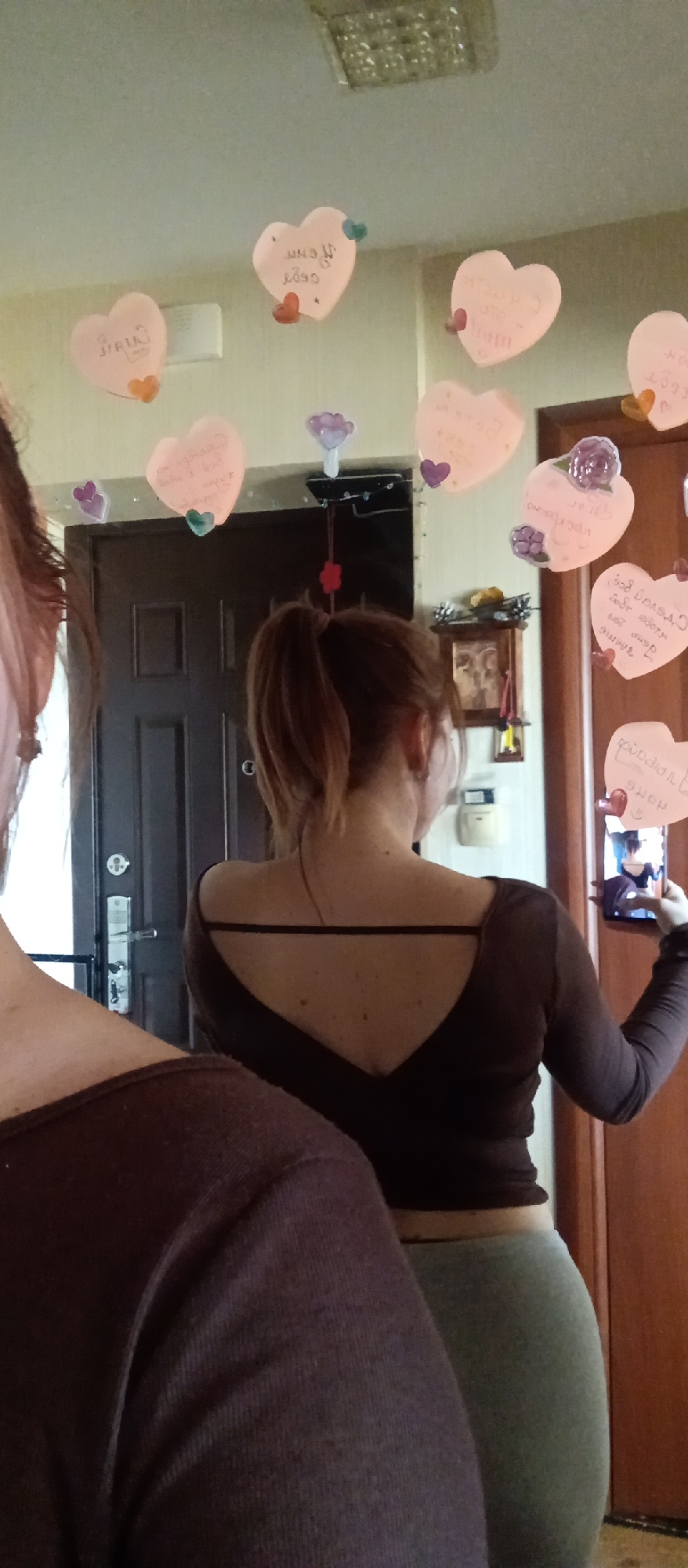 Two women are seen from behind in an indoor setting, likely a home, decorated with numerous heart-shaped notes. In the mid-ground, a woman with reddish-brown hair styled in a ponytail is wearing a dark brown long-sleeved top with a low V-shaped back and a horizontal strap, paired with light grey leggings. She is holding a smartphone up, capturing a mirror selfie, with her reflection visible on the phone screen. A portion of another woman with lighter hair is visible in the left foreground, also wearing a dark top.
The ceiling and walls are adorned with many light pink heart-shaped notes, some with small attached colorful plastic decorations like flowers and hearts. These notes feature handwritten text, which appears to be in Cyrillic script. Partially decipherable phrases include "С днем" (S dnem - with day), suggesting greetings like "С днем рождения!" (Happy Birthday!), and "Спасибо за" (Spasibo za - Thank you for...). This suggests a celebratory event such as a birthday or anniversary.
Behind the women, a dark brown door with a metallic handle and lock is visible, along with a small wall-mounted shrine or decorative box containing an image. The room is illuminated by an overhead ceiling light fixture.
