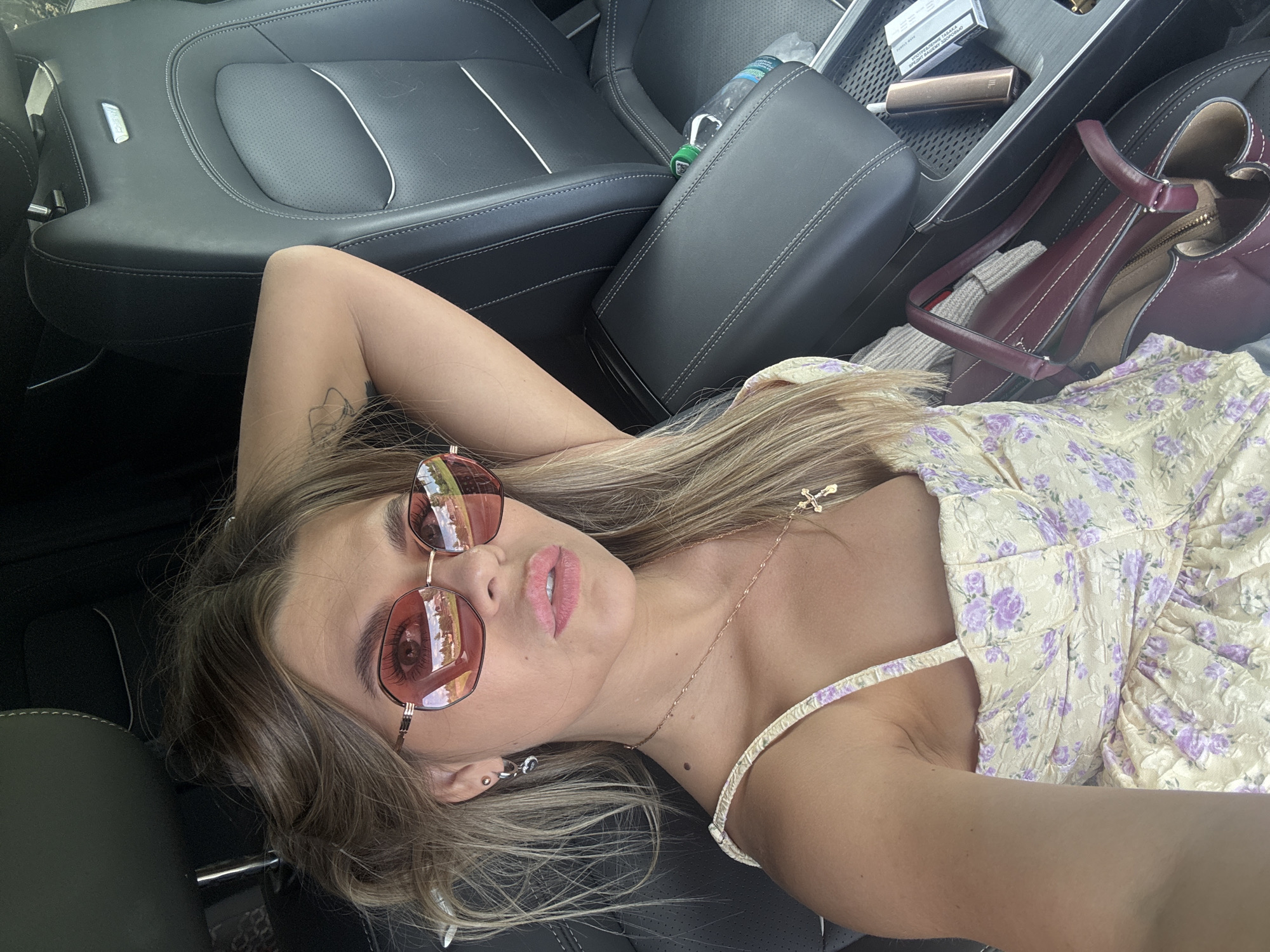 A young woman with blonde hair, wearing sunglasses and a light-colored floral dress, lies on the leather seats of a car. She is looking up at the camera with a relaxed expression. A small cross necklace is visible around her neck. The interior of the car is dark, with black leather seats. In the background, a purse and a bottle of water are visible. The scene appears to be set during the daytime, with bright lighting indicating good weather. The location is identified as Pastukhovo, Russia. There is text on a pack of cigarettes visible in the background, but it is not legible.