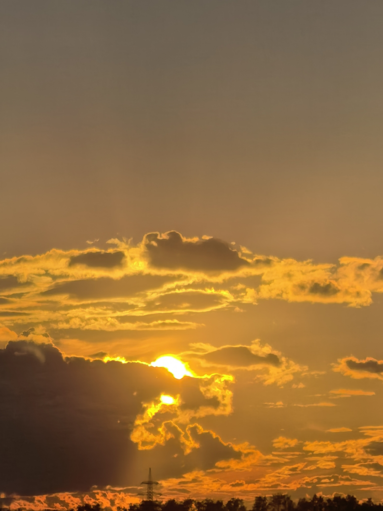 A vertical, medium shot captures a dramatic sunset over a silhouette of trees and a tall utility tower. The sky is a gradient of warm tones, ranging from a pale, muted orange at the top to a vibrant, fiery orange and yellow near the horizon. The sun, a brilliant white orb, peeks through and behind thick, golden-edged clouds, casting rays of light that illuminate the scene with a warm glow. Some darker, shadow-laden clouds are visible, creating depth and contrast. In the foreground, the dark silhouettes of trees line the bottom of the frame, interrupted by the stark, dark outline of a utility tower, suggesting an undeveloped or rural area. The overall impression is one of a serene yet powerful natural spectacle.