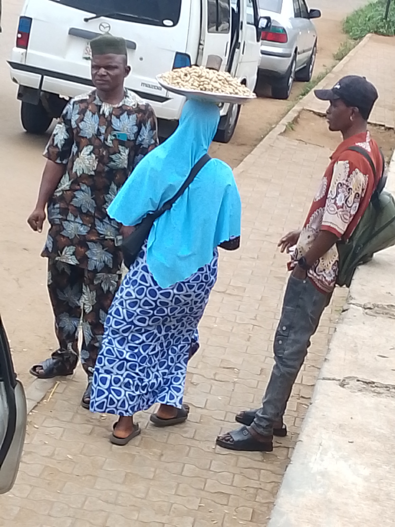 This image depicts a street scene in Oja Oba, Oko Erin, Nigeria. In the foreground, three people are visible. On the left, a man wearing a dark green cap and a patterned shirt stands looking to his right. He appears to be a vendor, given the tray of nuts balanced on the head of the person next to him. The central figure, wearing a bright blue headscarf and a patterned blue and white skirt, is carrying the tray of nuts. To the right, another man in a red and white patterned shirt, a black baseball cap, and dark pants stands facing left, with a backpack slung over his shoulder. He is holding a cigarette and appears to be interacting with the other individuals. The background includes a white van and a silver car parked on the street, suggesting an urban or semi-urban environment. The ground is paved with interlocking bricks. The lighting indicates it is likely daytime. The overall impression is of a casual street interaction, possibly involving commerce. There is no visible text in the image.