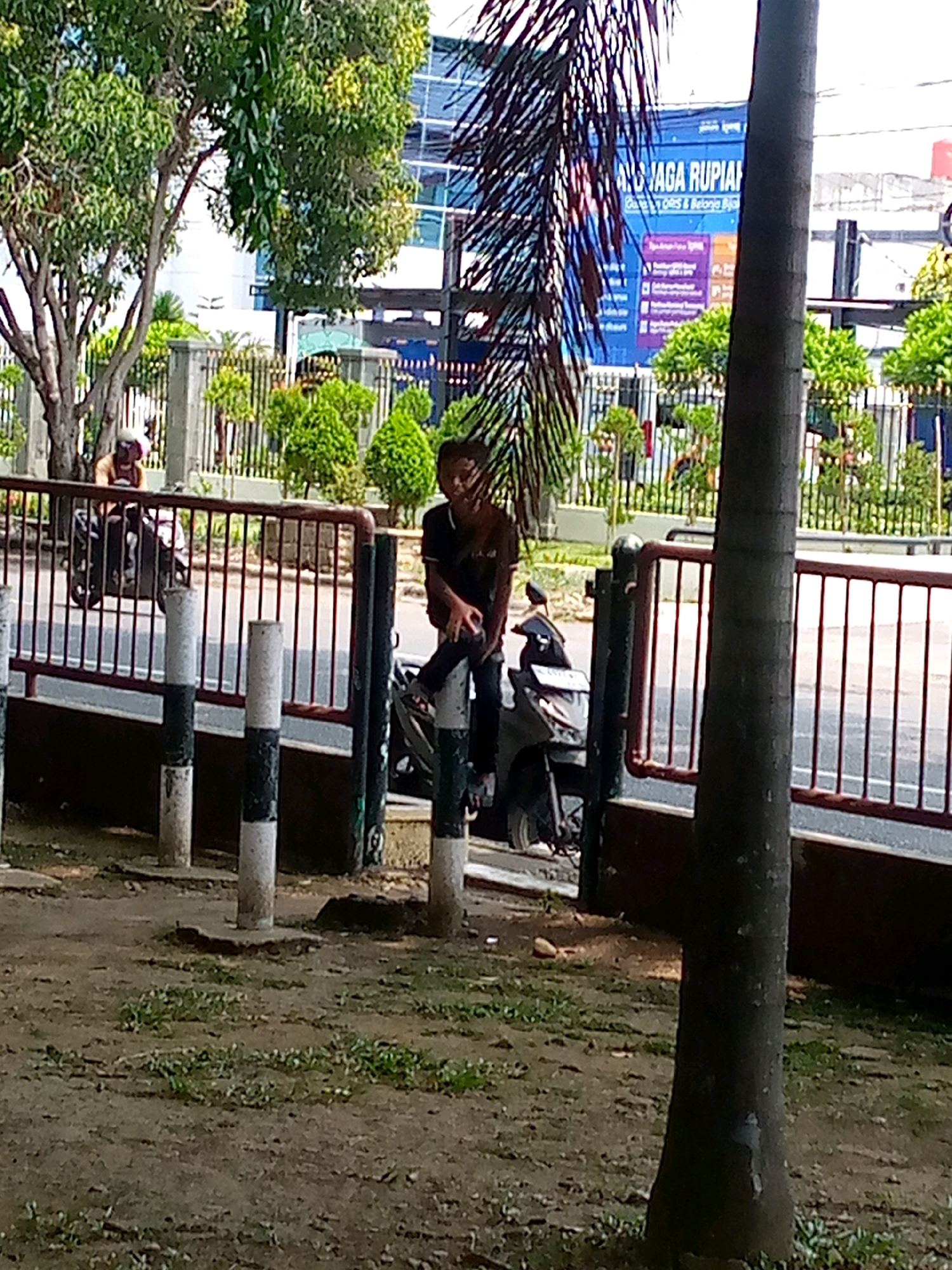 The image depicts a street scene in Lhokseumawe, Indonesia. In the foreground, a dirt path with sparse green vegetation leads towards a red metal fence. Several concrete posts, some with black and white markings, are positioned along the fence. Behind the fence, a person is seated on a black motorcycle. Further back, another person on a motorcycle is visible. A large tree with green foliage is present on the left side of the image. In the background, a modern building with blue windows stands prominently. A large blue banner with text is affixed to the building. The overall environment suggests an urban setting with a mix of natural and man-made elements.