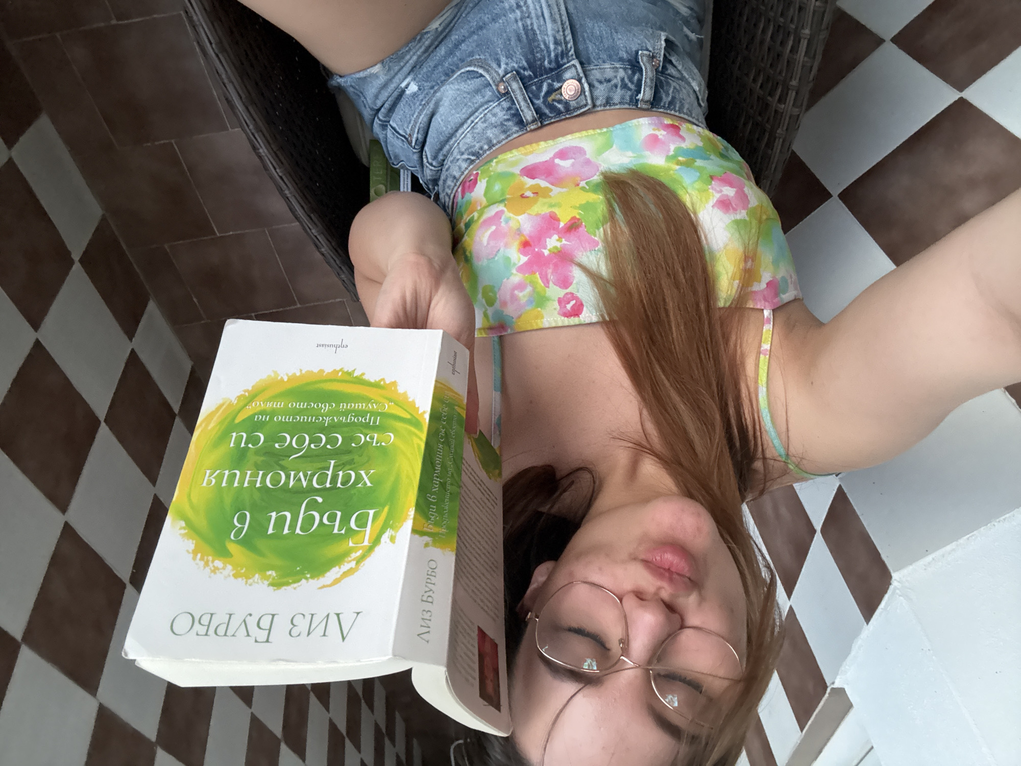A woman with red hair and glasses is reading a book titled "Бъди в хармония със себе си" (Be in Harmony with Yourself) by Liz Burbo. She's wearing denim shorts and a floral top and is sitting on a wicker chair. The background shows a tiled floor. It is likely this image was taken in Bulgaria, given the book is in Bulgarian. Given the tiles and wicker chair, it is probable that the location is a patio, likely at home or a cafe.