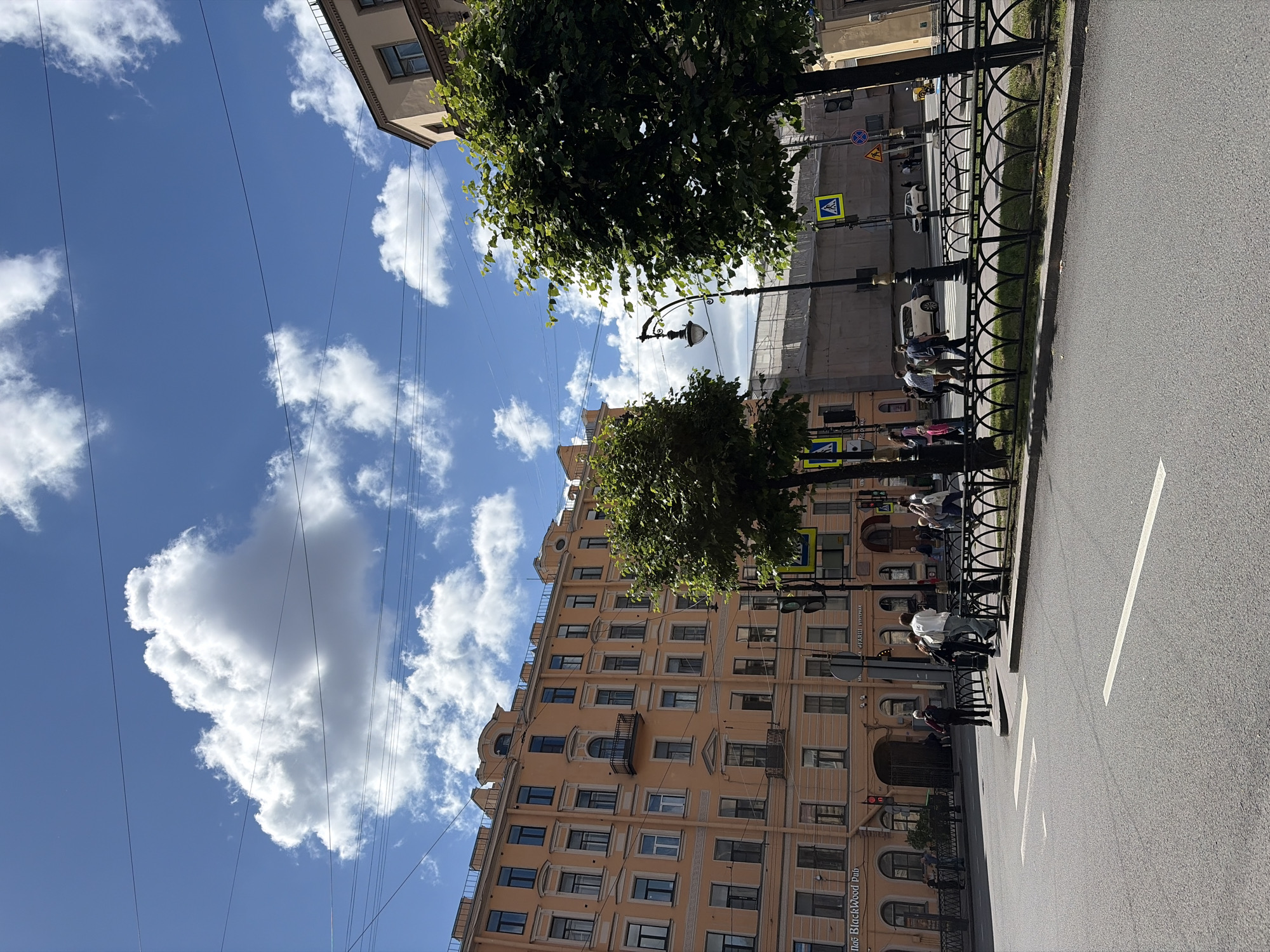 The image depicts a sunny day scene at a street intersection in Saint Petersburg, Russia, with a large peach-colored building dominating the left side of the frame. The building features multiple stories with numerous windows and decorative architectural elements, including balconies and a prominent arched entryway at street level. In front of the building, several people are walking on the sidewalk or crossing the street. A black metal fence with ornate, arched decorative elements runs along the sidewalk, separating it from the street. Two large, leafy green trees are visible, one to the right of the peach building and another larger one in the mid-ground to the right, partially obscuring a street lamp and a tall, modern building under construction or renovation behind it. The sky is a vibrant blue with scattered white clouds. Overhead power lines are visible against the sky. The street is asphalt with white lane markings. Traffic lights and pedestrian crossing signs are present at the intersection. A white car and a beige car are visible in the background.