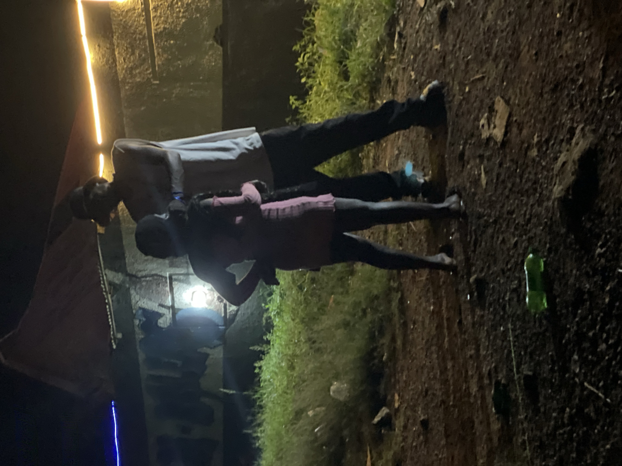 A man and a girl stand outside a building at night. The man is taller than the girl, and they appear to be in an intimate embrace. They are both dressed in casual clothing. The building behind them is lit by yellow and blue lights. The ground in front of the building is covered with dirt and grass. There is a green plastic bottle on the ground. The overall scene suggests a casual gathering or social interaction in an outdoor setting. The specific location in Durimi, Nigeria, cannot be determined from the image alone.