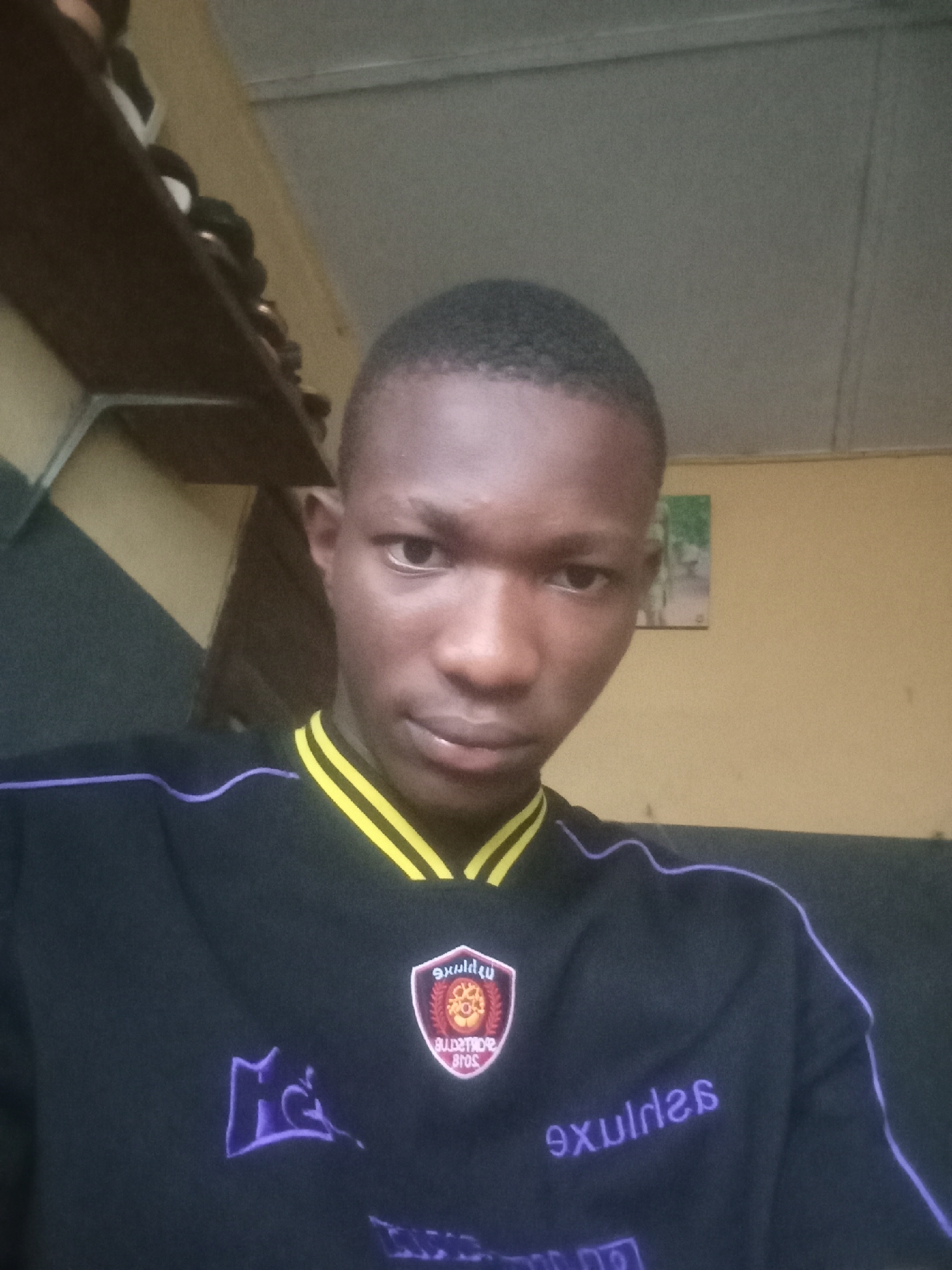 A close-up, head-and-shoulders selfie shows a young Black male with short hair wearing a black jersey with yellow trim around the collar and purple trim detailing on the sleeves. The jersey has a crest on the left chest and a stylized logo on the right chest. The background appears to be an indoor setting with a shelf on the left and a yellow wall with a framed picture on the right. The lighting is dim, and the image quality is moderate.