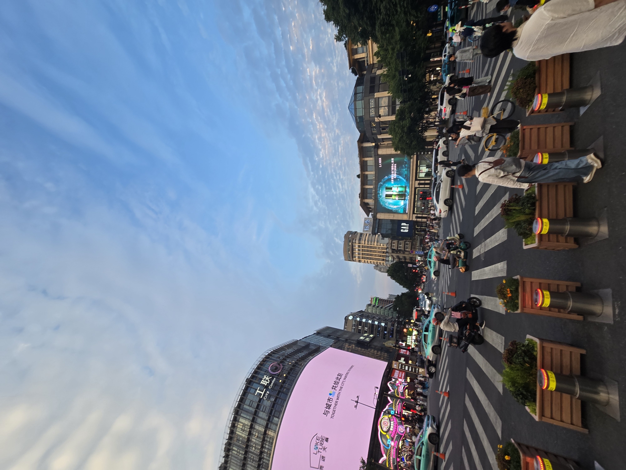 The image captures a bustling street scene in Shangcheng District, China, likely during the late afternoon or early evening. The sky is a mix of blue with wispy white clouds. Dominating the left side of the frame is a large, curved building with a massive pink LED screen displaying text in Chinese and English that reads "TOGETHER WITH THE CITY, PAINTING TRENDS". Below this, a neon-lit facade suggests a shopping area or entertainment venue.
The street is an active intersection with multiple lanes of traffic. Cars, including several green sedans, and motorcycles are visible. Pedestrians are crossing a wide crosswalk marked with bold white stripes. Some individuals are pushing bicycles, and others are walking or standing.
On the right side of the image, more buildings line the street, featuring commercial signage. One prominent display shows "GAP". Further down the street, a tall, modern building with illuminated windows stands out. Green trees are visible along the sidewalks, adding a touch of nature to the urban landscape. The foreground features planters with greenery and decorative bollards with blinking red lights. The overall atmosphere is one of urban activity and commerce.