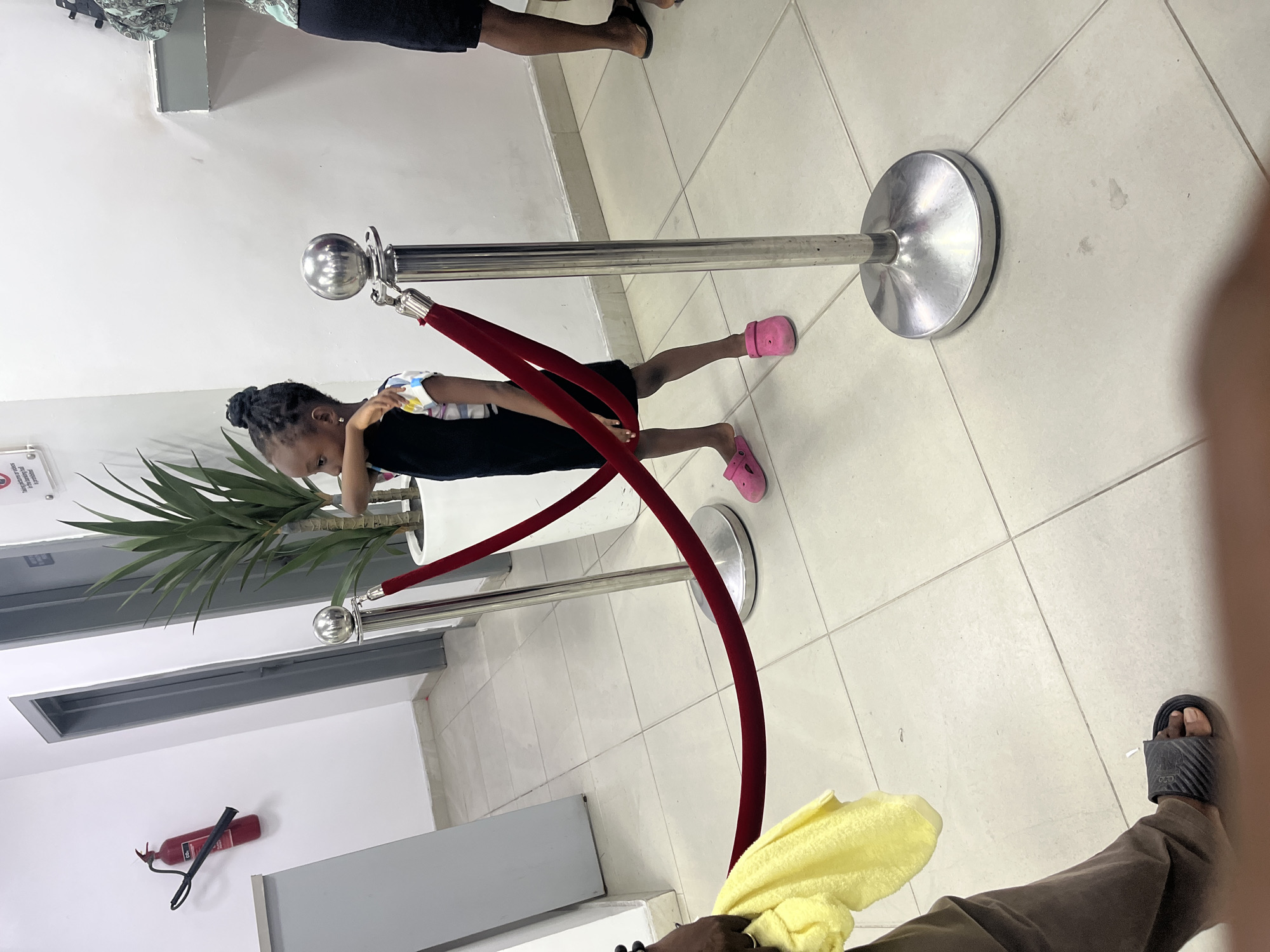 A young girl with dark hair braided on top of her head is lying on her stomach, face down, under a red velvet rope attached to two chrome stanchions in Ado, Nigeria. She is wearing a black dress with a white, floral-patterned yoke. Her legs are stretched out towards the right side of the frame, and she has on pink Crocs. The stanchions are positioned on a light-colored tiled floor.
Behind the girl and to the left, there is a potted plant. On the wall, there is a fire extinguisher. The legs of two other people, one wearing black shorts and the other wearing brown pants and sandals, are visible on either side of the frame. A yellow towel is also partially visible in the lower right corner.