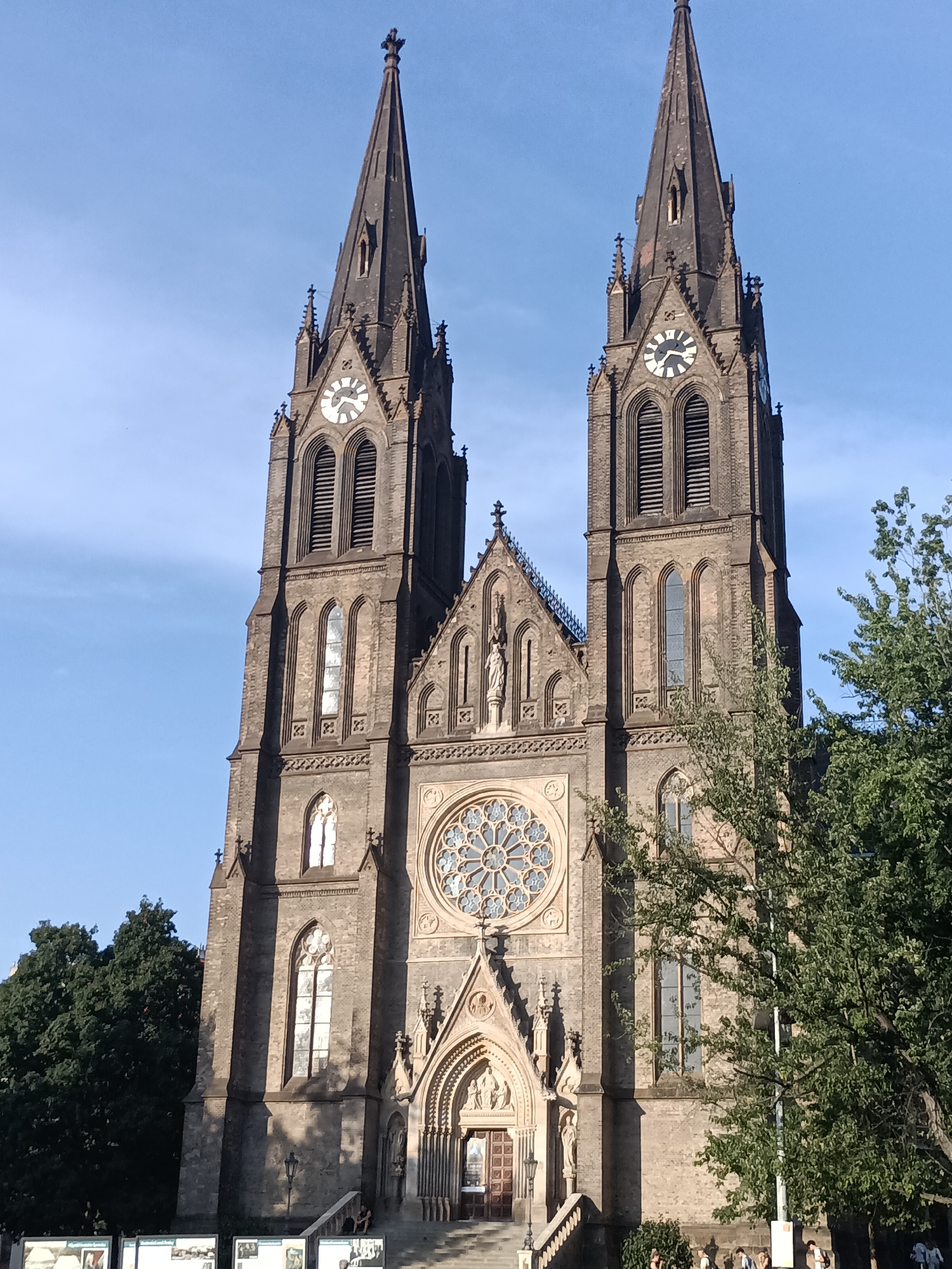 The image shows the Basilica of St. Peter and St. Paul, located in Vyšehrad, Prague, Czechia. The church is a prominent Gothic Revival building with two tall spires, each topped with a cross. Both spires feature clocks near their apexes, with the left spire's clock showing approximately 3:50 and the right spire's clock indicating approximately 3:45. The facade of the church is made of brick and features arched windows, decorative stonework, and a large rose window above the entrance. A statue of a figure, possibly a saint, is visible on the facade between the two spires. The entrance to the church is a large, ornate archway with a wooden door.
In the foreground, a set of stairs leads up to the entrance of the church. Several people are visible on the stairs and at the base of the church, some appearing to be tourists. Green trees, including a large tree in the right foreground, frame the lower part of the church. The sky is clear and blue, indicating a sunny day. The overall scene is one of a grand architectural landmark under a bright sky.
