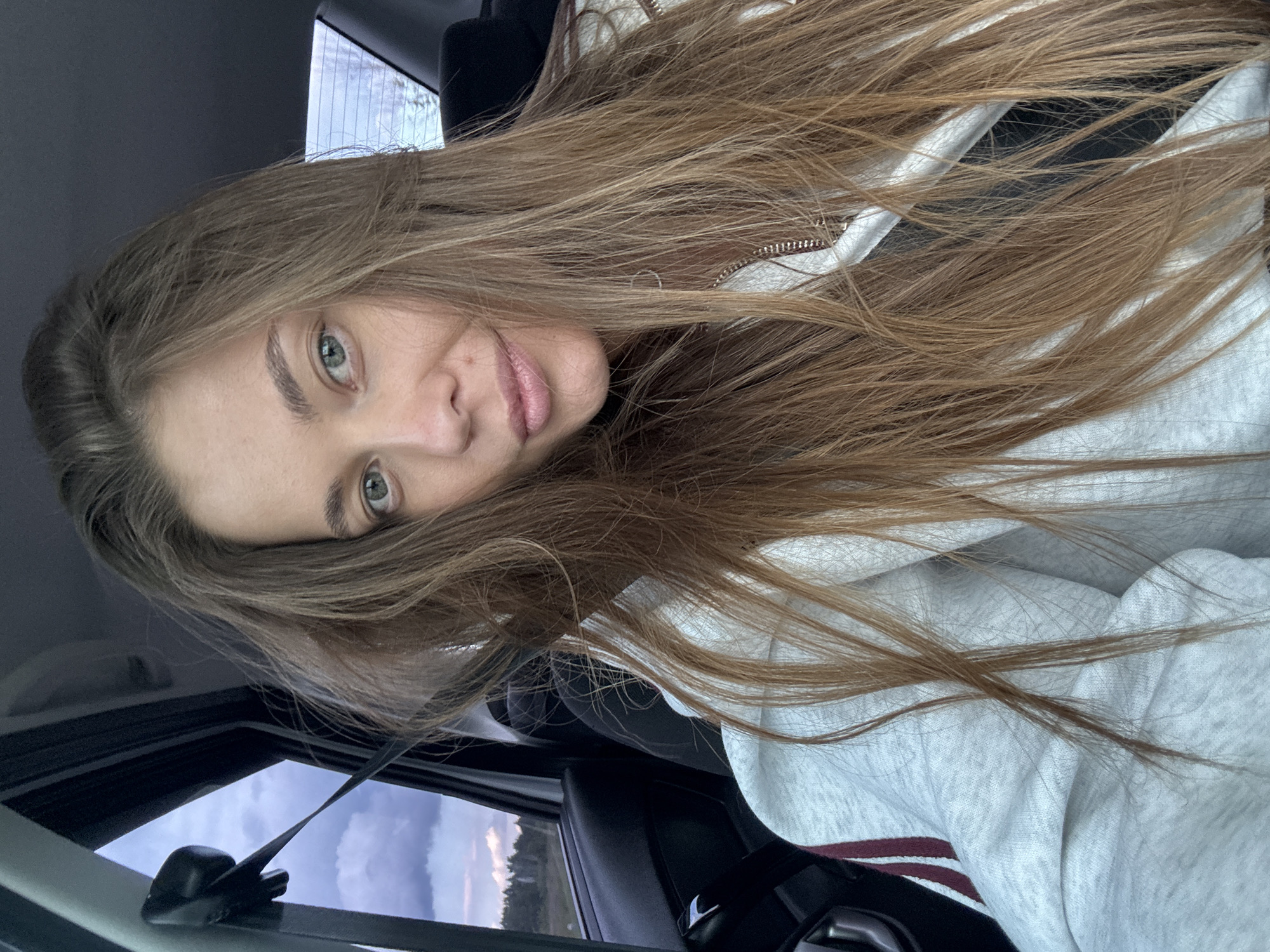 A young woman with long, light brown hair is captured in a selfie from inside a car. She is wearing a light gray hooded sweatshirt with dark red stripes on the sleeves. Her eyes are open and looking towards the camera, and her lips are slightly parted. The background shows the interior of the car, including a seatbelt, and through the window, a view of a cloudy sky over a treeline and a rural landscape. The image does not contain any elements that would suggest the location in the city of Бикбарда, Russia.