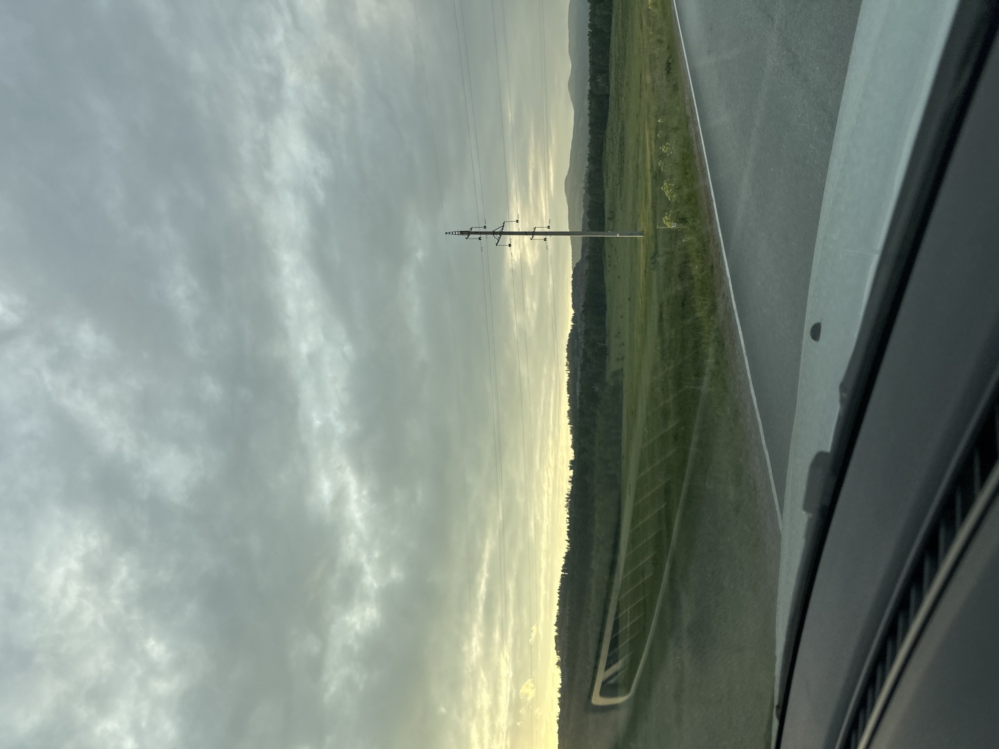 The image, taken from inside a vehicle, shows a landscape scene under a cloudy sky. A power line runs vertically across the left of the frame. Adjacent to the power line is a green field with a drainage ditch running parallel to the road. A line of trees separates the field from distant hills. Part of the vehicle interior is visible in the lower right corner. The image appears to be rotated counter-clockwise. The location is unknown.