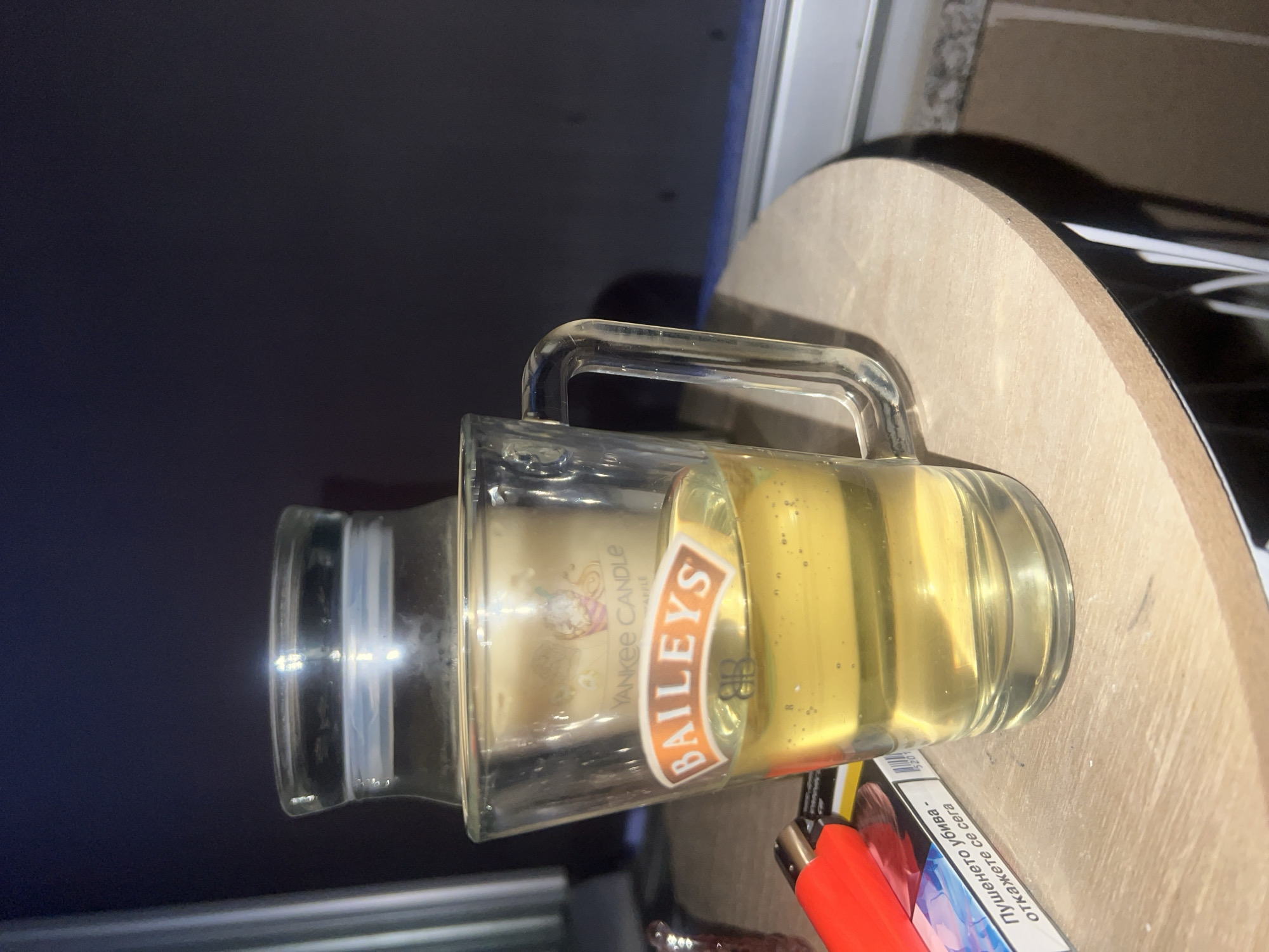 The image shows a glass with Baileys liqueur on a wooden table, with a lit cigarette pack and a lighter in the foreground. Behind the glass, a Yankee Candle is visible. The scene appears to be indoors, possibly on a balcony, with a window in the background. The lighting suggests it is nighttime. There are no people or animals in the image. The location is not Plovdiv, Bulgaria.