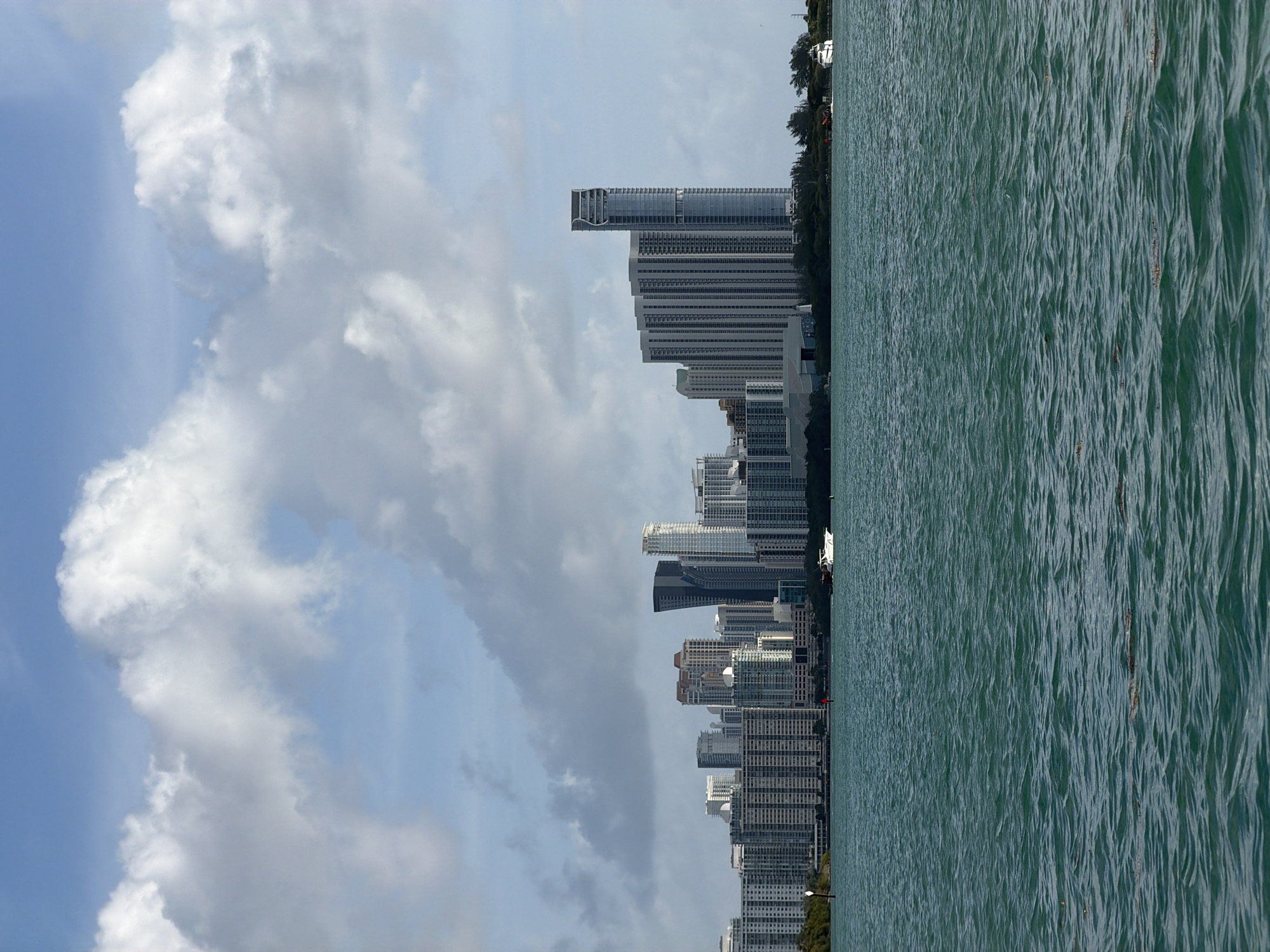 The image shows a view of Miami, Florida, United States of America. The cityscape is prominent, with numerous tall buildings and skyscrapers lining the coastline. To the left of the buildings, there are formations of white clouds in a blue sky. To the right, the scene is dominated by water with visible ripples and patterns. There is no visible indication of it being Bal Harbour Village.
