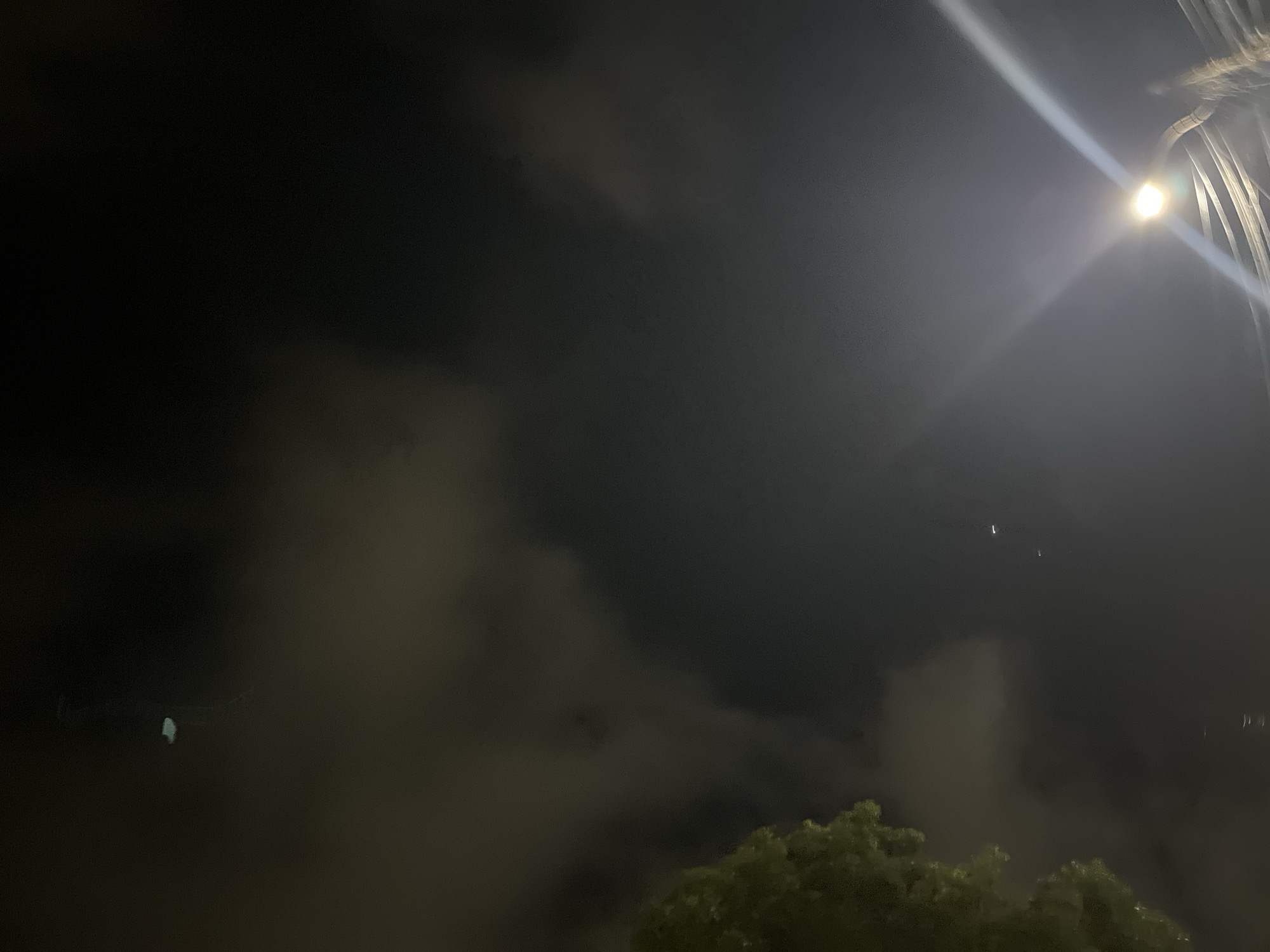 The image depicts a night sky with clouds and a street light. In the upper left quadrant, a small, bright object is visible against a dark cloud. A portion of a tree with green leaves is visible on the far left side of the image. A bright street light illuminates the lower right quadrant, casting light beams across the frame. Several faint lights are scattered in the middle right of the frame. The scene appears to be an outdoor urban environment at night, potentially in Santa Ana, Venezuela, given the context provided.