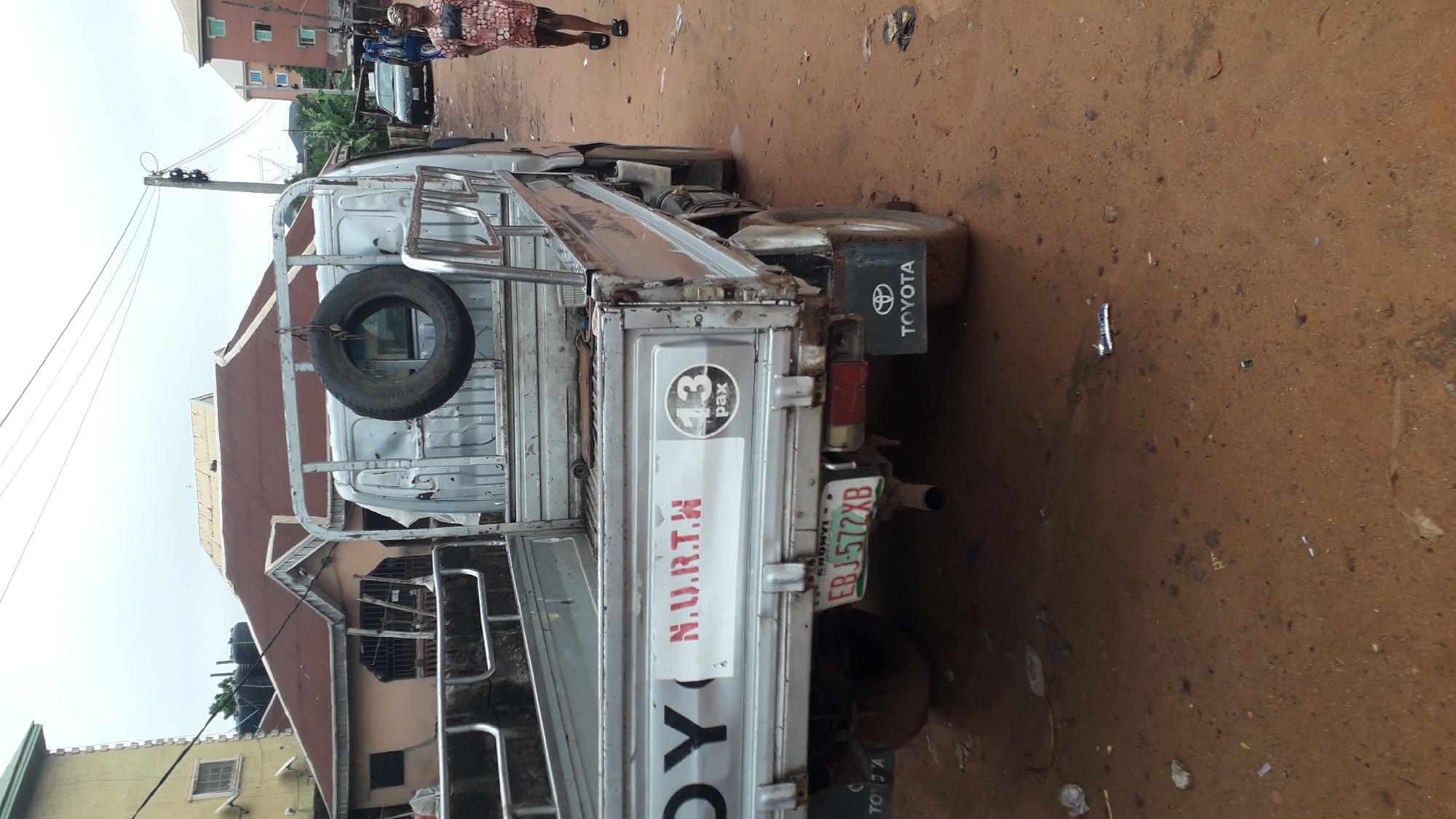 The image depicts the rear of a silver Toyota pick-up truck, located in Nkpor, Nigeria. The truck bed is exposed with metal framework. A spare tire is secured to the back of the cab. The tailgate displays "N.UR.T.H." in red lettering, and a "13 pax" sticker is present. The license plate reads "EBJ-572XB." Two people are partially visible in the upper portion of the image, standing on a dirt road. Buildings and overhead power lines are in the background. The ground is reddish-brown dirt, scattered with trash.