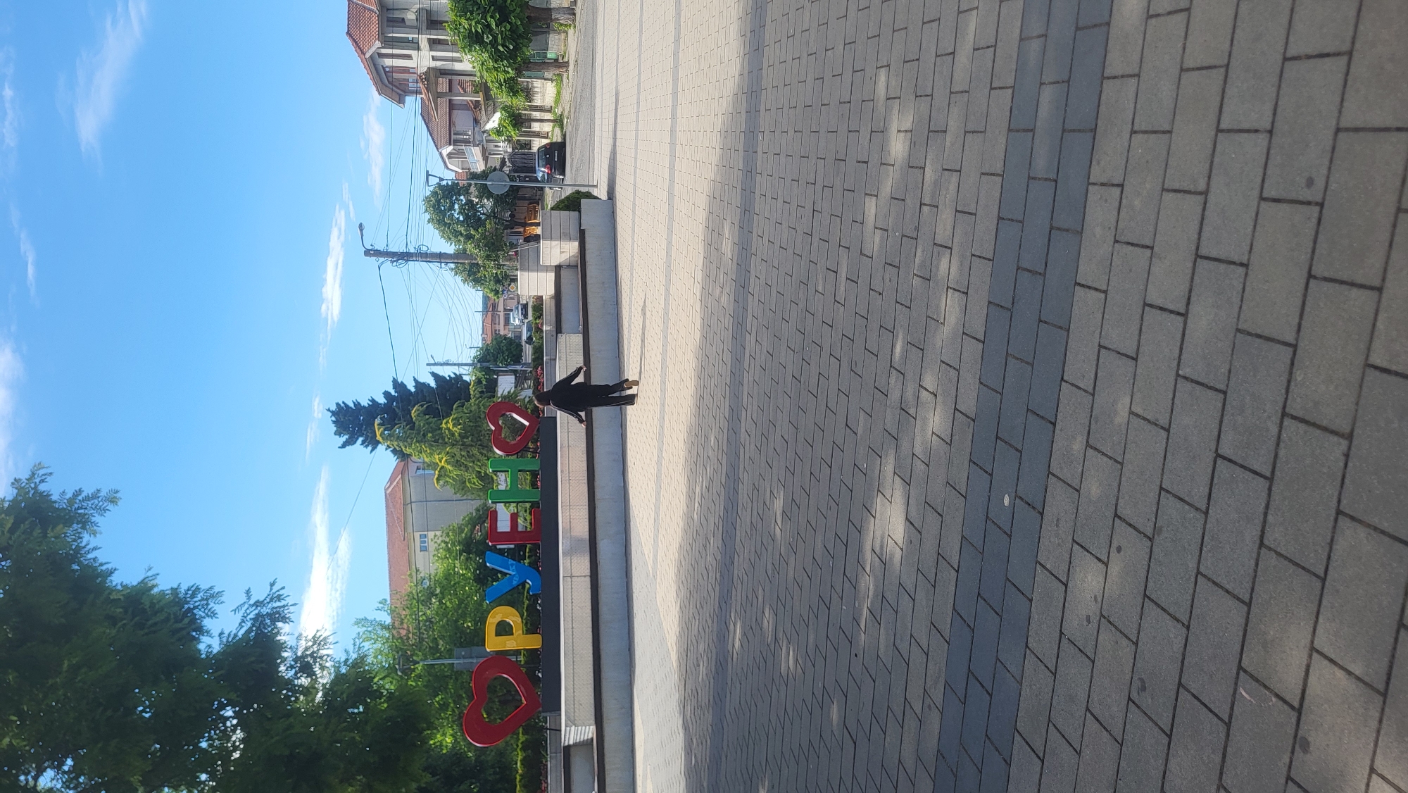 The image depicts a bright, sunny day in Ruen, Bulgaria, with a clear blue sky. In the foreground, a paved area made of gray rectangular bricks is visible, with shadows cast from overhead trees. Further back, a large, colorful sign with the word "РУЕН" (Ruen) written in block letters is prominently displayed. The letters are a mix of red hearts and yellow, blue, and green letters. A person, dressed in dark clothing, is walking away from the camera towards the sign. In the background, there are trees, residential buildings, and a street with a few cars. The overall scene suggests a public square or a town center.