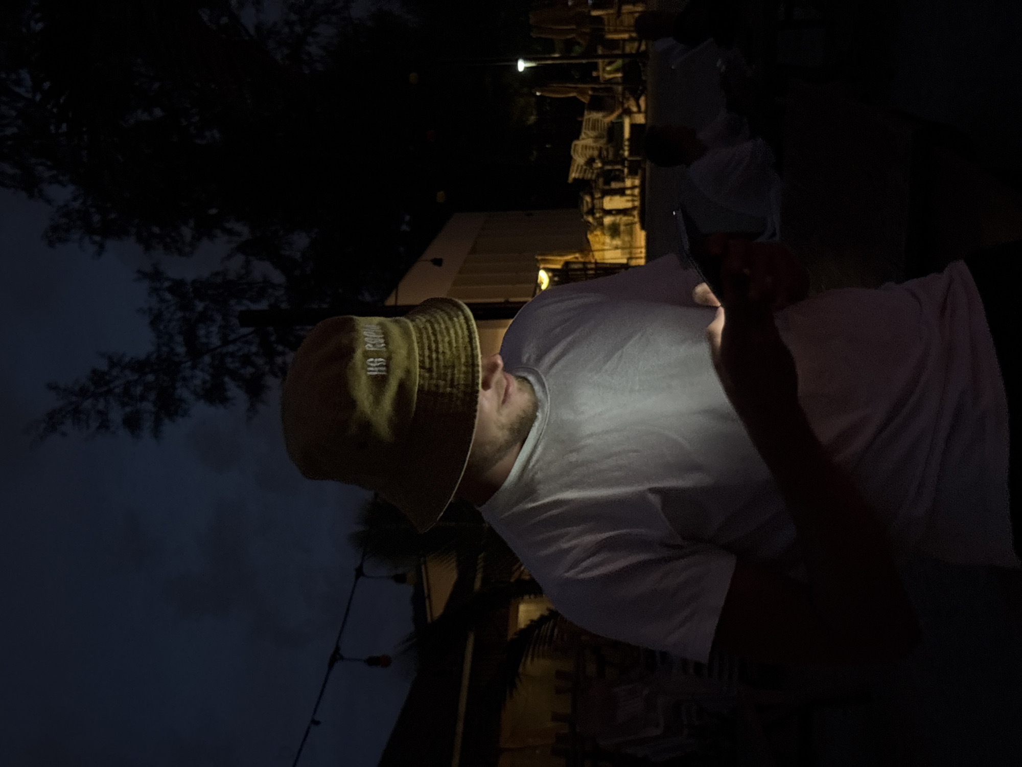A person wearing a light-colored t-shirt and a bucket hat is looking at their phone. The phone's screen illuminates the person's face, which is partially obscured by the hat. The background is dark, suggesting it is nighttime. In the background, there are glimpses of lights and what appear to be outdoor chairs and tables, hinting at a restaurant or seating area. The person is the central focus of the image, with their attention directed downwards at the device. The lighting on the person's face is a key element, creating a dramatic contrast with the surrounding darkness. The image captures a moment of personal engagement with technology in a dimly lit outdoor environment. The visible text on the hat reads "US ROCK". The time of day is night.
