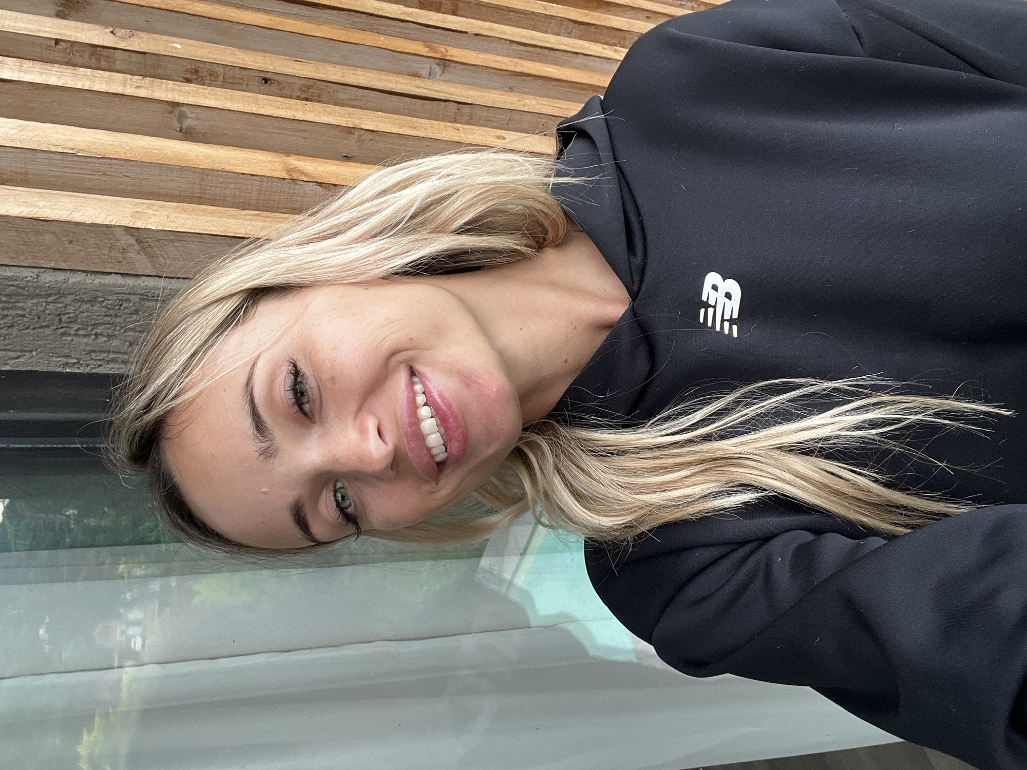 A blonde woman smiles at the camera. She has green eyes and her hair is styled in loose waves. She is wearing a black hoodie with a white New Balance logo on the left chest. The background consists of a sheer curtain and a wooden wall. The image appears to have been taken outdoors, possibly on a balcony, as evidenced by the presence of greenery reflected in the window. The scene does not depict any specific location in the city of Tsodoreti, Georgia.