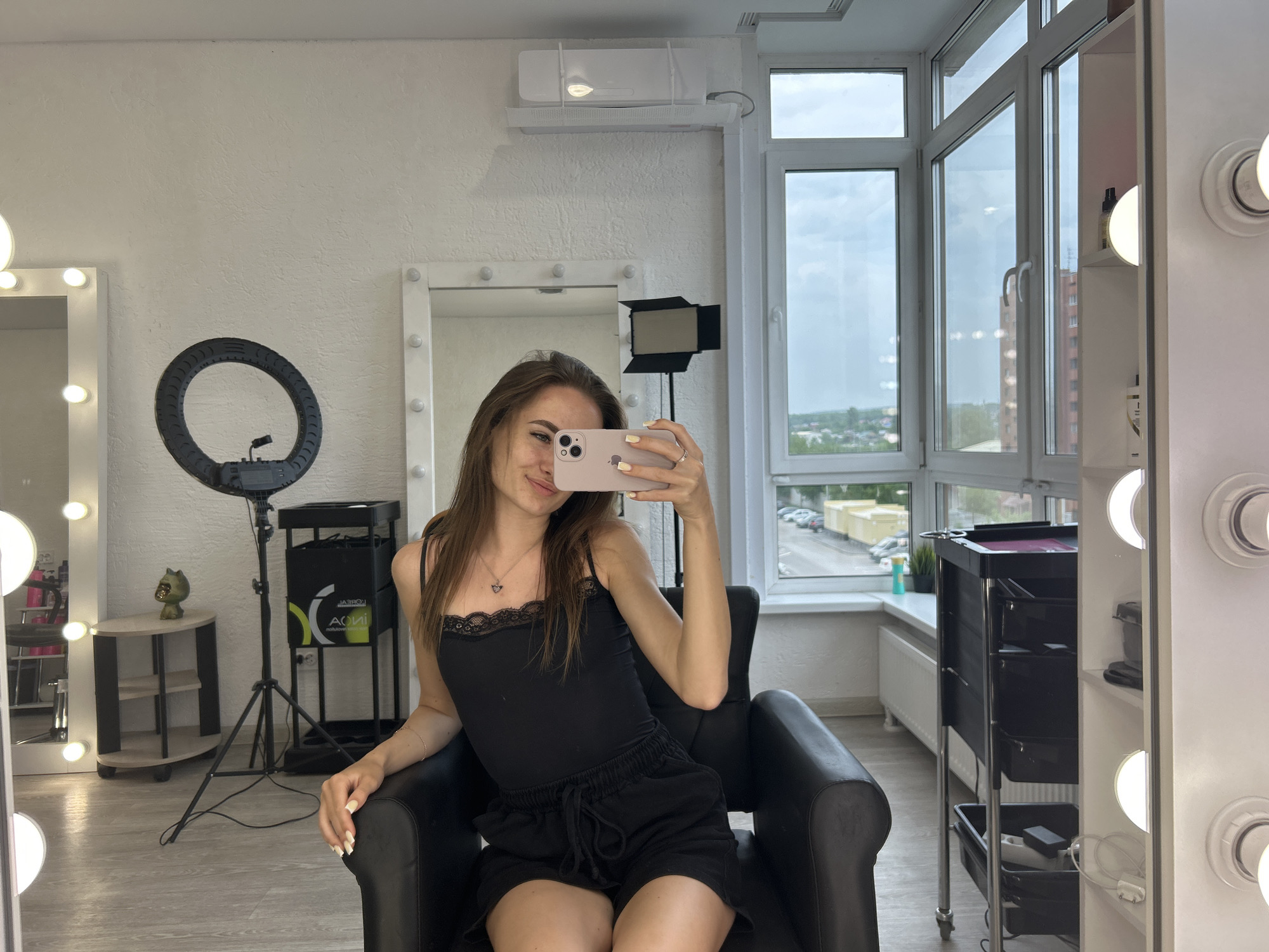 In an indoor studio setting with large windows overlooking a city, a young woman with long brown hair is seated in a black leather chair, taking a selfie with a pink smartphone. She is wearing a black lace-trimmed camisole and black shorts. Behind her, a ring light stands on a tripod, and to her right, a multi-tiered trolley holds various items. The studio features a white textured wall and a light-colored wooden floor. Natural light streams in from the windows, illuminating the space.