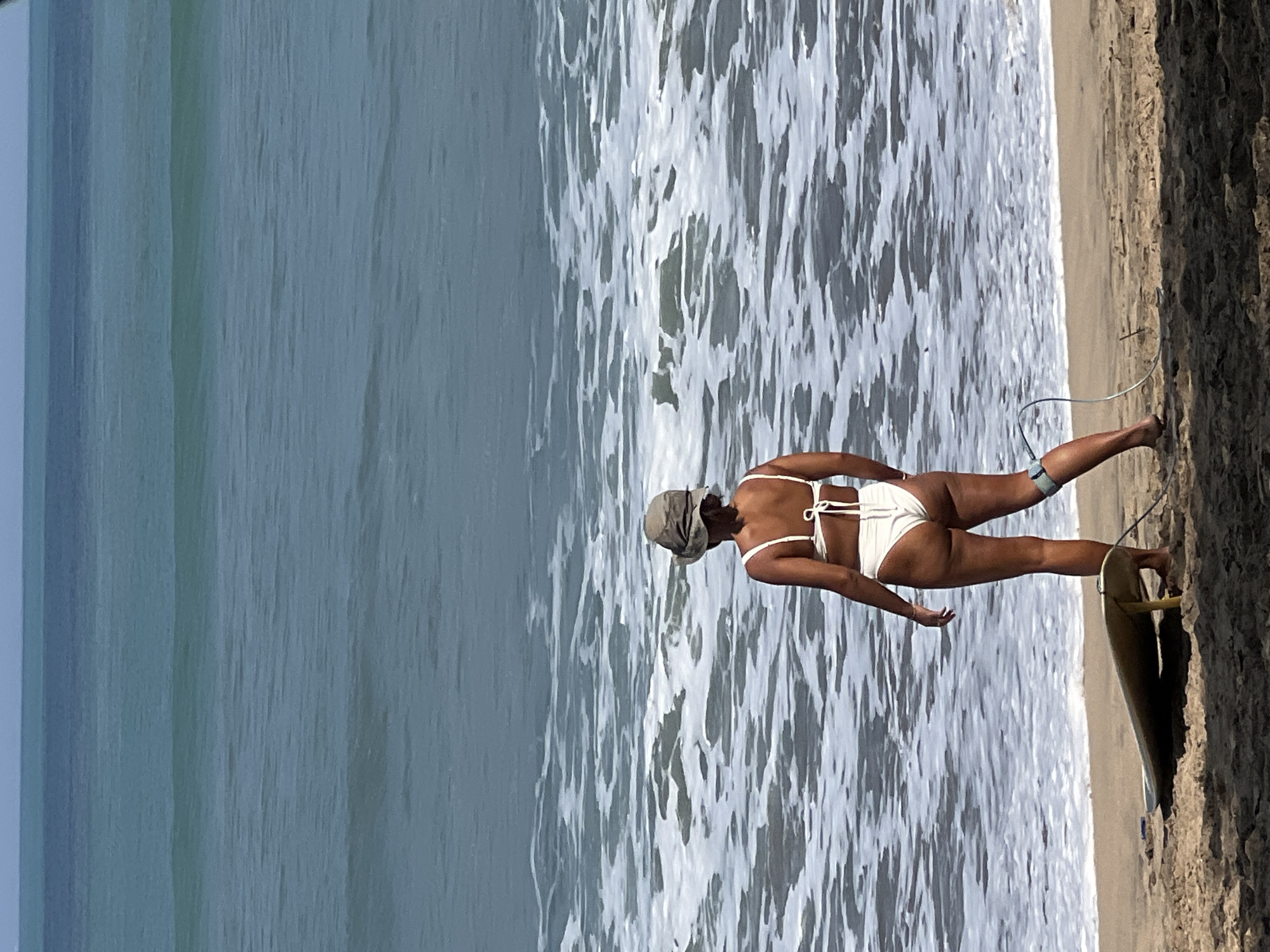A woman in a white bikini and a bucket hat stands on a sandy beach, facing the ocean in Kuta, Indonesia. A surfboard is propped up on the sand next to her. The ocean is blue with white waves breaking on the shore. The sky is clear and bright.