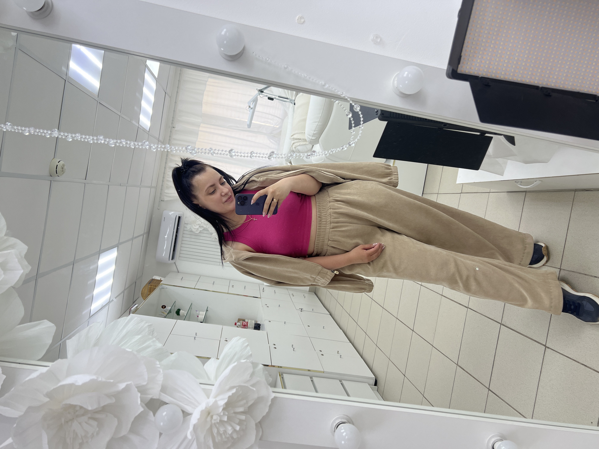 A young woman with dark hair tied in a ponytail, wearing a pink tank top, a beige corduroy jacket, and beige corduroy pants, takes a selfie in a full-length mirror. She is also wearing dark blue and white sneakers. In the reflection, she is standing in a brightly lit room with white tiled floors and white walls. To the left of the woman, large white paper flowers are attached to the wall. In the background, a row of white cabinets with gold handles is visible, along with a red bottle of Coca-Cola on one of the shelves. A mirror with lights around its frame is the central focus of the image. The room appears to be a salon or beauty studio, with a massage bed and a lamp visible in the far right of the mirror's reflection. The woman is holding a dark-colored smartphone in her right hand. The overall impression is that of a personal moment captured in a clean, modern environment.