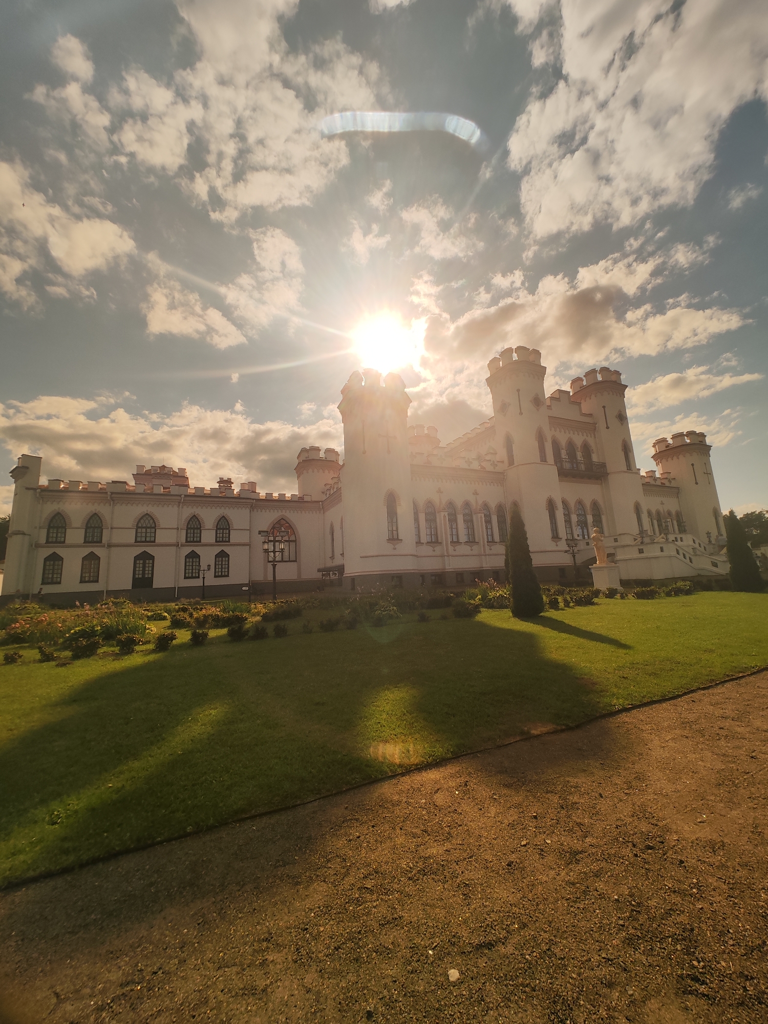 The image shows a white, neo-gothic style castle, possibly a manor house, with numerous turrets and decorative elements. The castle is situated on a grassy lawn with flowerbeds in the foreground. A gravel path runs along the right side of the image. The sun is shining brightly from behind a partially cloudy sky, creating lens flare and casting long shadows across the lawn. The castle appears to be located in an open, park-like setting. The context provided places the location in the village of Kvasivichy, Kvasivichy Rural Council, Belarus.