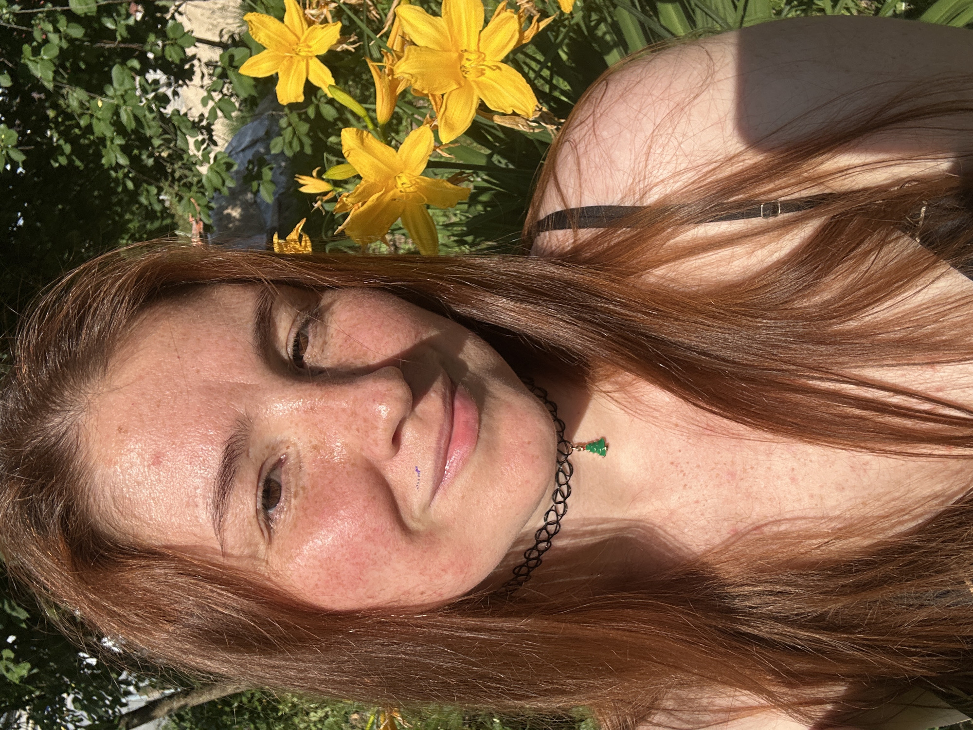 A close-up selfie of a young woman with long, auburn hair is captured outdoors on a sunny day. She is positioned in front of a dense background of green foliage and bright yellow lilies. The woman is looking directly at the camera with her right eye closed and her left eye partially open. She has a light complexion with numerous freckles across her face, cheeks, and décolletage. A black choker necklace adorns her neck, featuring a small, green, Christmas tree-shaped charm. Her shoulders and upper chest are visible, revealing a hint of a black garment. The lighting suggests the sun is positioned to her right, casting subtle shadows across her face and hair. The overall scene evokes a sense of casual outdoor leisure. The specific location within Nizhny Novgorod, Russia cannot be determined from the visual information provided.
