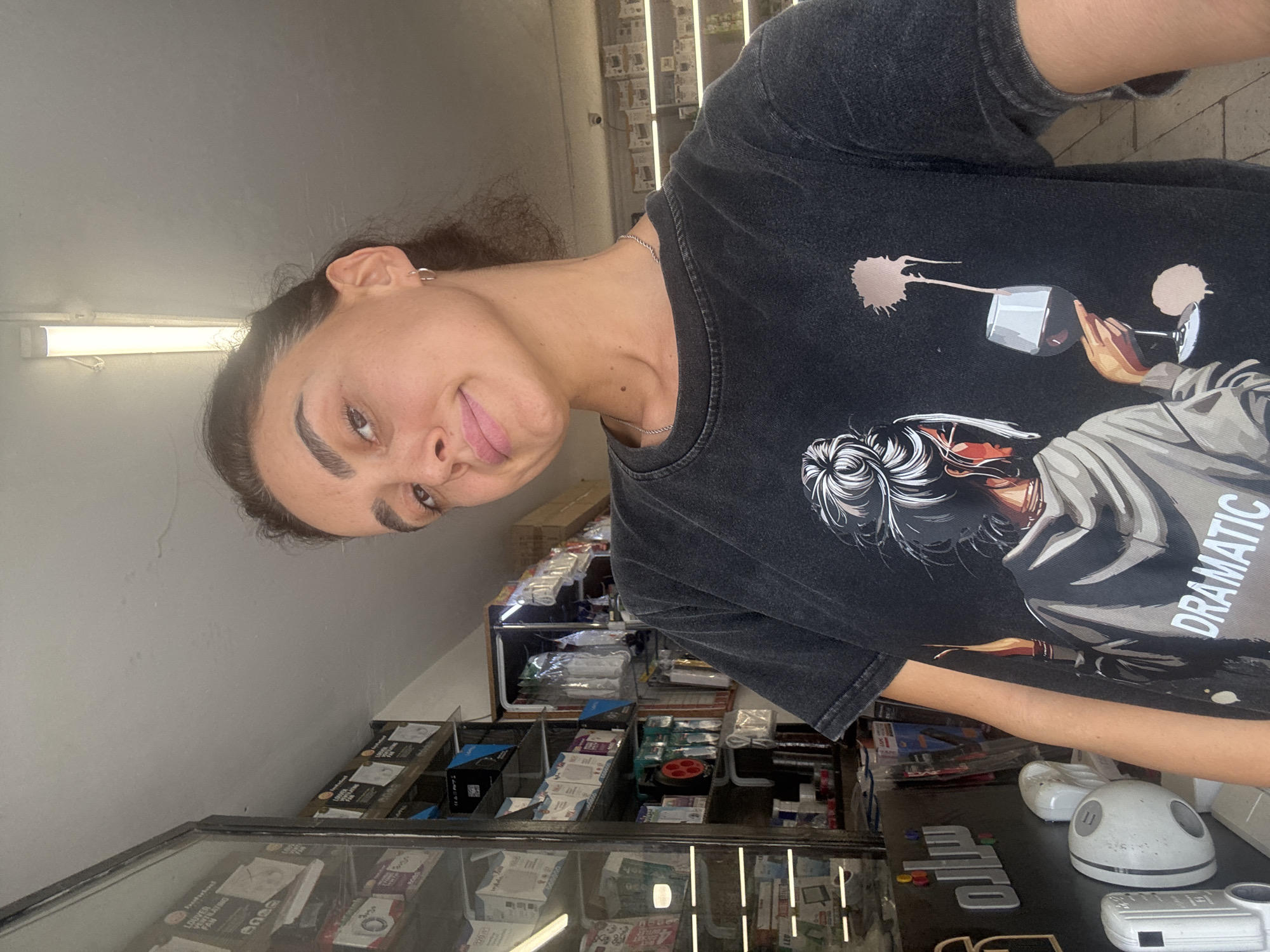A young woman with dark hair pulled back in a bun, wearing a dark t-shirt with a graphic print, stands in what appears to be a shop. The shop is stocked with shelves filled with various items, possibly electronics or hardware, with boxes and packaging visible. A glass display case is on the left side of the frame, containing more merchandise. The woman is looking towards the camera, and the lighting suggests it is daytime. The background shows more shelves and displays, indicating a retail environment. The overall scene is a casual selfie taken inside a shop.