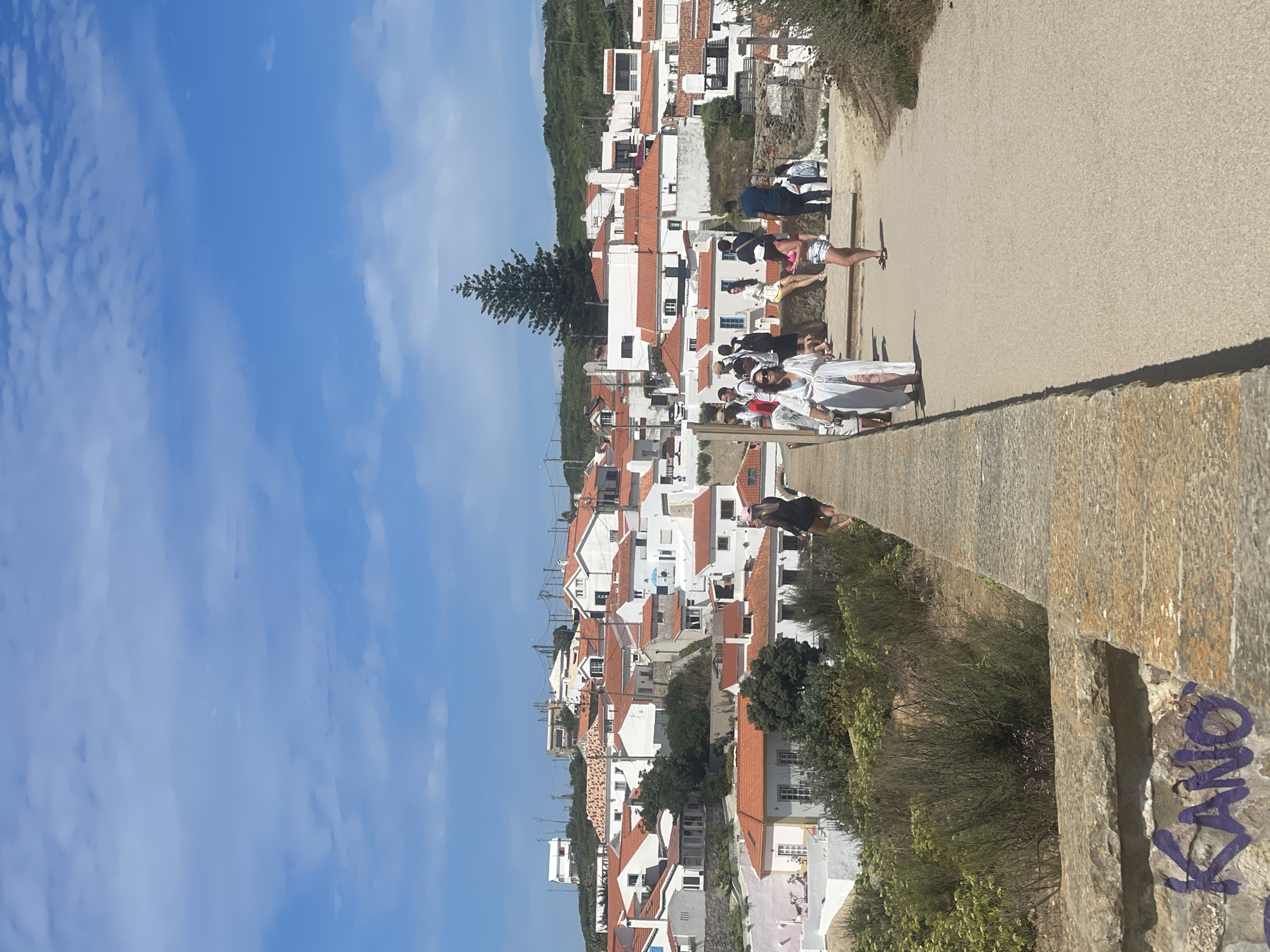 The image captures a view of the town of Colares, Portugal, with a cluster of white buildings featuring orange-tiled roofs nestled on a hillside. A pathway with a stone wall borders the foreground, on which purple graffiti spelling "KANO" is visible. Several people are scattered along the pathway and on a raised area overlooking the town. In the mid-ground, a group of individuals are seen walking and standing, some appearing to be tourists. A tall pine tree stands prominently against the blue sky, dotted with white clouds. The overall scene depicts a sunny day in a picturesque coastal town.