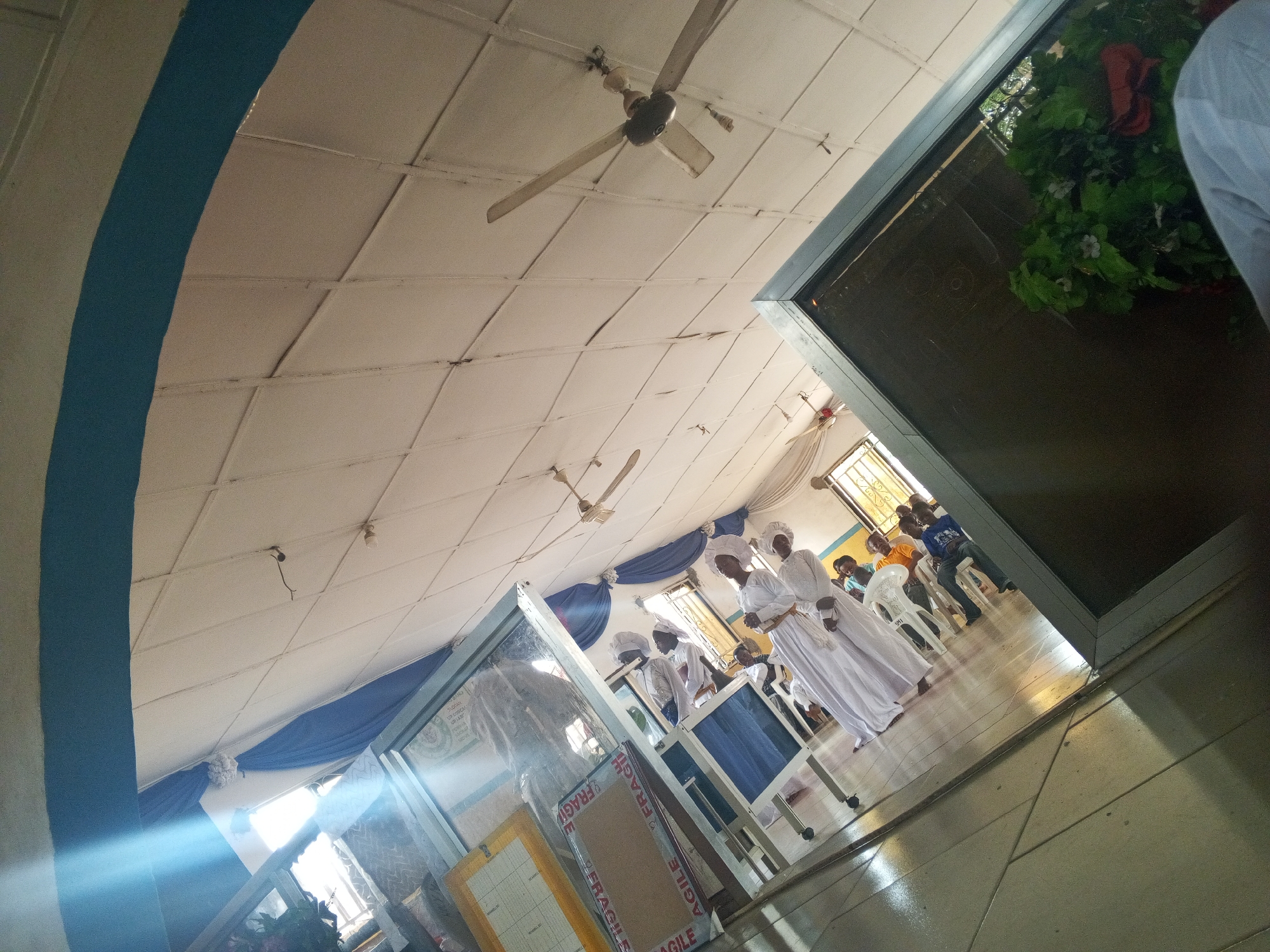 The photograph captures a church interior in Lokoja, Nigeria. Several individuals dressed in white garments and head coverings are positioned near what appears to be a railing or partition. Other people are seated in white plastic chairs in the background. The room has a white ceiling with visible ceiling fans. Blue drapery adorns part of the space. In the foreground, "FRAGILE" is visible on stacked boxes. A window reflects the interior. The perspective of the photograph is at an angle looking slightly upwards.