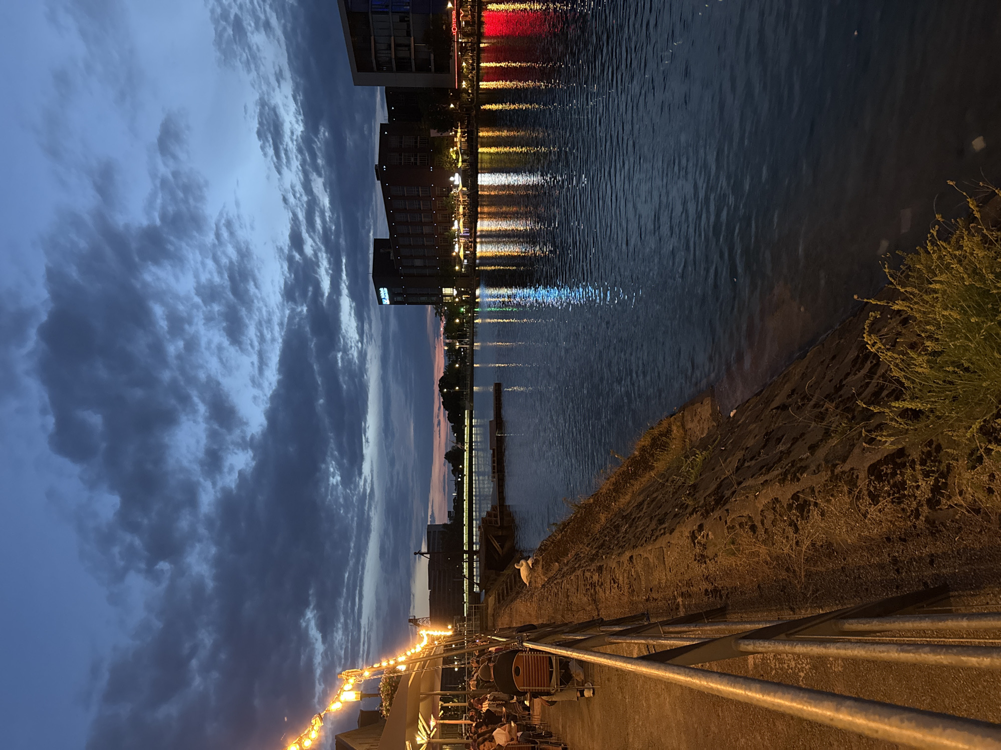 The image depicts a twilight scene at the Innenhafen (inner harbor) in Duisburg, Germany. The sky is a deep blue with scattered dark clouds, transitioning to a hint of orange and pink on the horizon. On the left side of the frame, an outdoor seating area with tables and chairs is visible, illuminated by string lights. Several people are seated at the tables. A metal railing runs along the edge of the walkway.
The water of the harbor occupies the right side of the image, reflecting the lights from the buildings on the opposite bank. These reflections are in various colors, including white, yellow, blue, and red. A stone embankment lines the harbor, with a swan resting near its edge.
In the background, across the water, are several modern buildings. One building, labeled "Penta Hotel," is prominent. Other buildings show illuminated windows and signs, suggesting businesses or residences. A bridge or walkway structure spans across the water in the distance. The overall atmosphere is calm and tranquil, characteristic of an evening by the water.