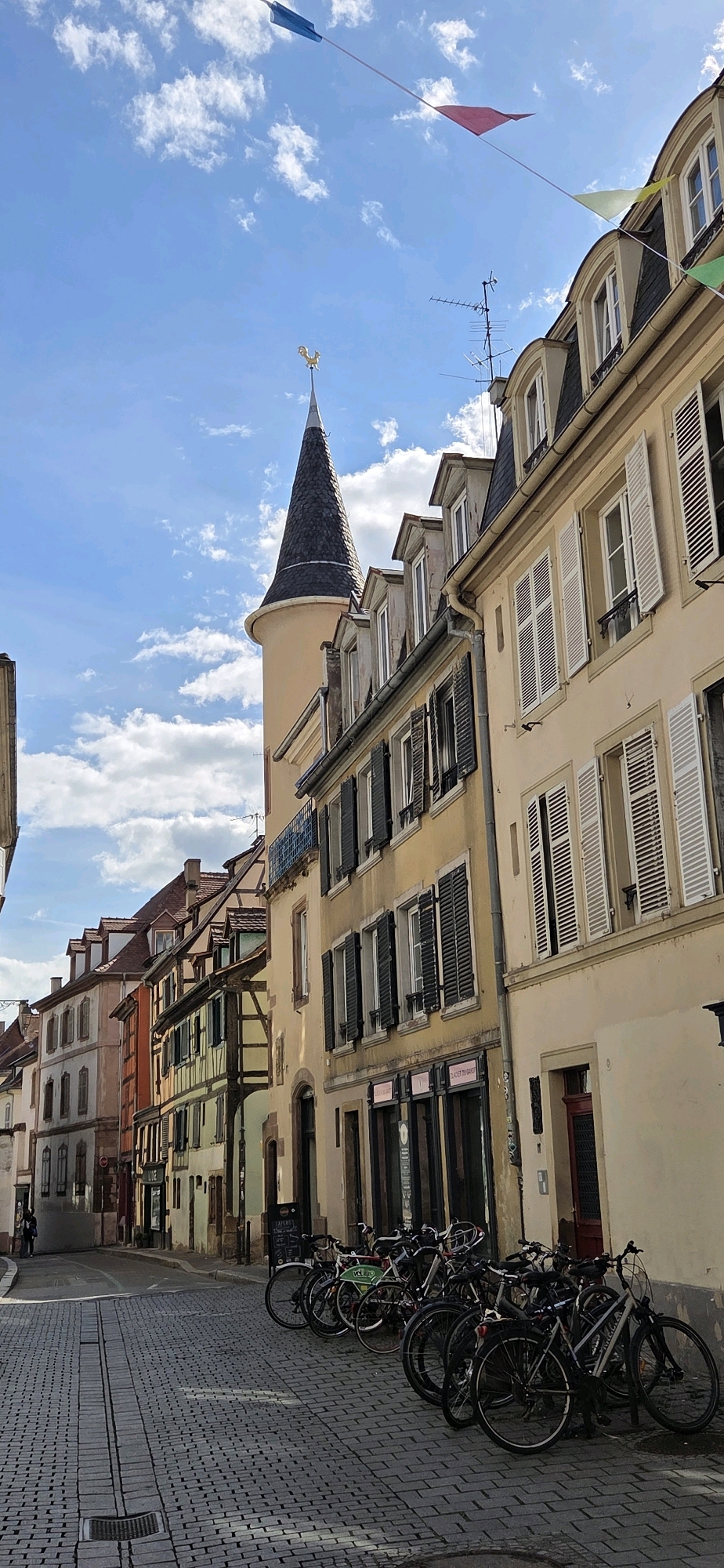 This image depicts a cobblestone street in Strasbourg, France, lined with historic European architecture. The buildings feature traditional multi-story facades with pitched roofs and dormer windows. Many of the windows are adorned with closed shutters, some in dark wood and others in white. A conical tower, topped with a gold rooster weather vane, is visible on the left side of the frame, adding a whimsical architectural element. Colorful triangular banners are strung across the bright blue sky, which is dotted with white clouds.
In the foreground, several bicycles are parked along the side of the street, leaning against each other. The cobblestone pavement is laid in a uniform pattern. The scene suggests a quiet, picturesque urban environment, likely a historic district within Strasbourg. The lighting indicates it is daytime, with sunlight casting distinct shadows on the street. The overall impression is one of old-world charm and a relaxed atmosphere.