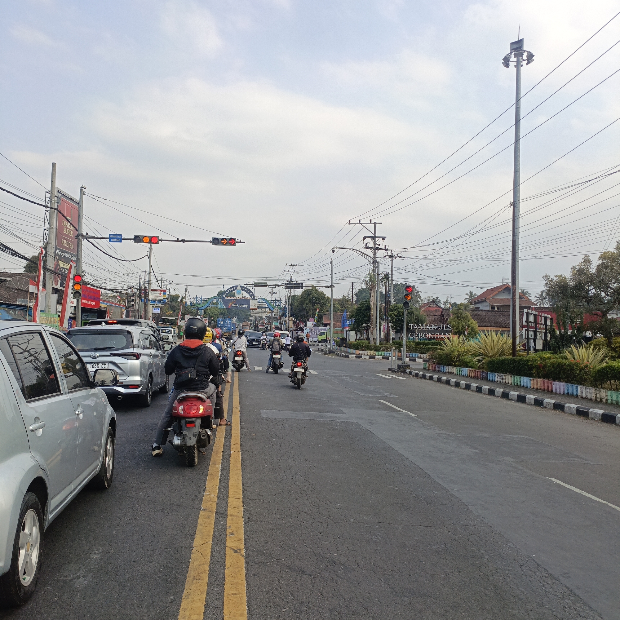 The image captures a traffic scene in Salatiga, Indonesia. In the foreground, a silver car is stopped on the left side of the road, while a red scooter with a rider wearing a black helmet and an orange-trimmed jacket is to its right, also stopped. Further ahead, several other scooters and cars are visible in traffic. Overhead, power lines crisscross the sky, and traffic lights are present. On the right side of the road, a tall light pole stands with multiple lamps. Beyond that, a sign reads "TAMAN JLS CEBONGAN." A decorative, colorful border lines the edge of the road. In the background, an arched gateway with the text "SELAMAT JALAN KOTA SALATIGA" is visible, indicating the city limits. The sky is overcast with scattered clouds. The overall scene depicts a typical urban traffic congestion.