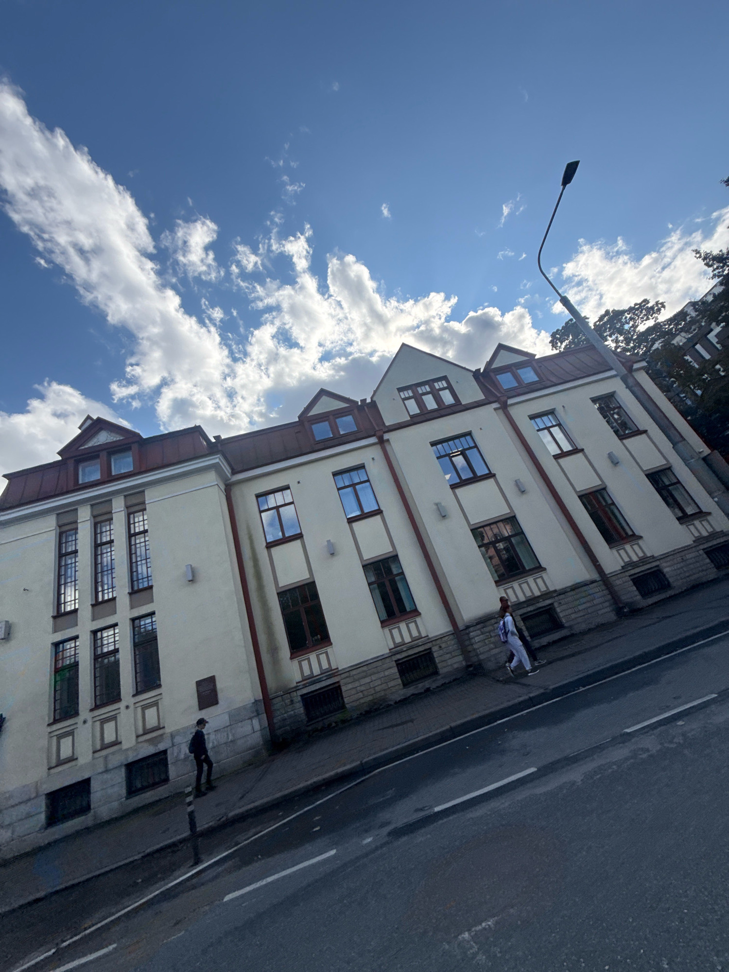 The image depicts a street scene in Tallinn, Estonia, featuring a large, light-colored building with a reddish-brown roof. The architecture suggests a historical or institutional building, with multiple windows and a symmetrical facade. The building is situated alongside a paved road, with a sidewalk running in front of it.
Two individuals are visible on the sidewalk. One person, wearing dark clothing and a cap, is walking away from the camera. Another person, in lighter colored athletic wear, is walking towards the camera, accompanied by a second person, partially obscured.
The sky above is blue with scattered white clouds, indicating daytime. A street lamp with a distinctive curved arm is positioned to the right of the building, silhouetted against the sky. The overall impression is a relatively calm urban environment on a cloudy day. The image has a slight tilt, giving it a dynamic perspective.