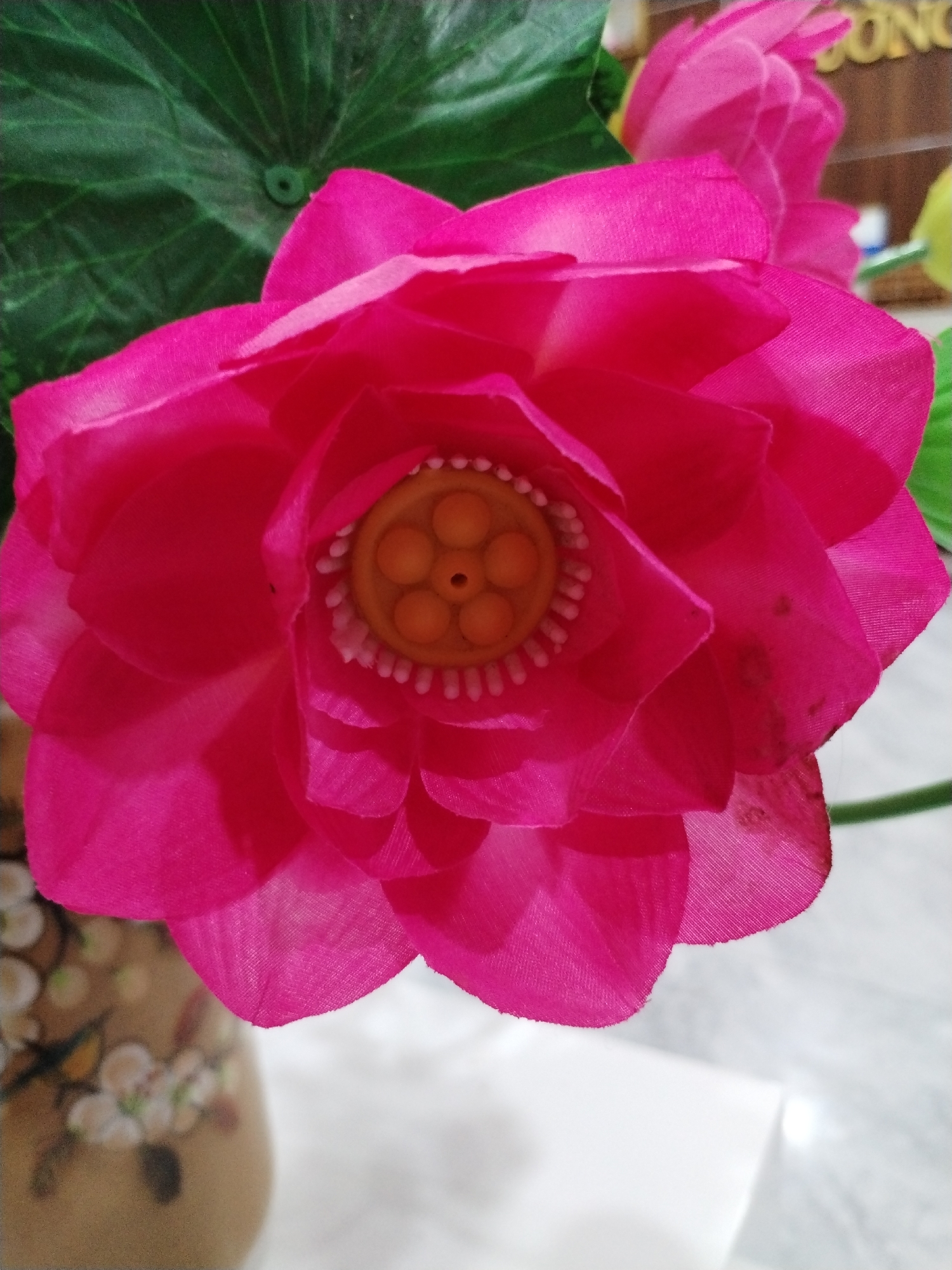 A close-up shot features a vibrant, fuchsia-pink artificial lotus flower dominating the foreground, angled slightly from the bottom left. The flower, likely made of fabric, displays multiple layers of petals with a slight sheen. Its intricate center is composed of a yellow-orange, disc-like structure with five raised, circular segments and a central dimple, surrounded by a ring of small, white, bead-like details.
Behind the prominent flower, another, less defined pink artificial flower is visible towards the top right. A large, dark green artificial leaf with prominent veins and a small, raised circular element at its center is seen in the upper left background.
On the bottom left, the rim and part of the body of a ceramic or wooden vase are visible. It is light brown or beige with a subtle, lighter floral pattern of what appear to be white or light-colored blossoms. The background beyond the flowers and vase is softly blurred, showing a light-colored, possibly white or grey, surface, likely a table or countertop. In the top right corner, a small portion of a sign with partially visible text, possibly "SONG" or "CONG," can be discerned. The overall setting appears to be indoors, showcasing a decorative arrangement in Bac Nha Trang Ward, Vietnam. No people or specific activities are depicted. The lighting suggests an indoor environment without direct sunlight.