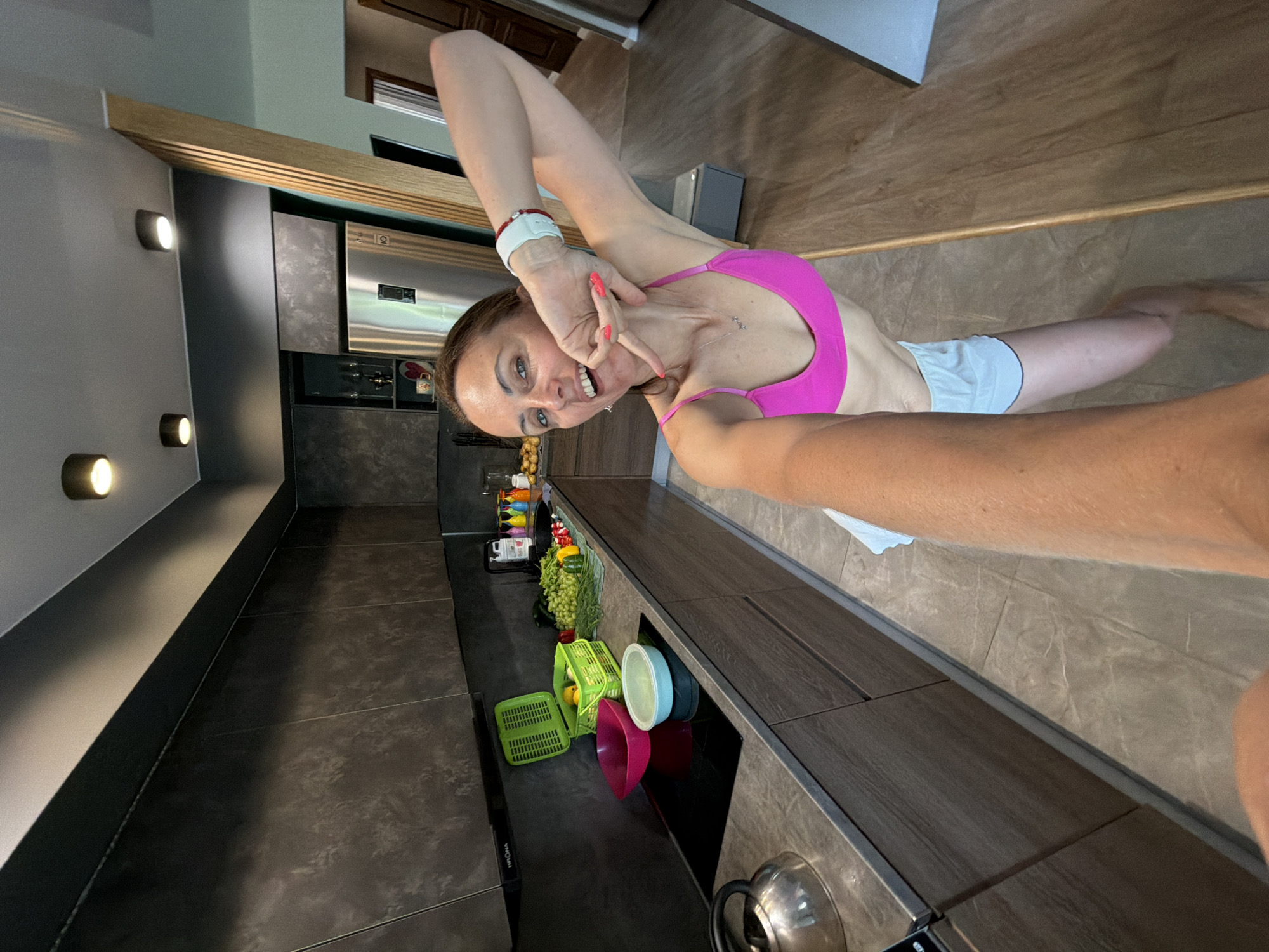 A woman with auburn hair is taking a selfie in a modern kitchen. She is wearing a bright pink sports bra and white shorts. Her left arm is extended, holding the camera, and her right arm is bent upwards, with her hand near her face. She is smiling and looking at the camera.
The kitchen features dark grey cabinets and countertops, with stainless steel appliances, including a large refrigerator. On the countertop, there are various items, including a green plastic basket filled with fruit, grapes, peppers, and other vegetables, as well as bowls and a kettle. The floor is tiled with a light brown, marble-like pattern. The overall aesthetic is sleek and contemporary.
The image does not contain any information that would place the location in Birobidzhan, Russia.