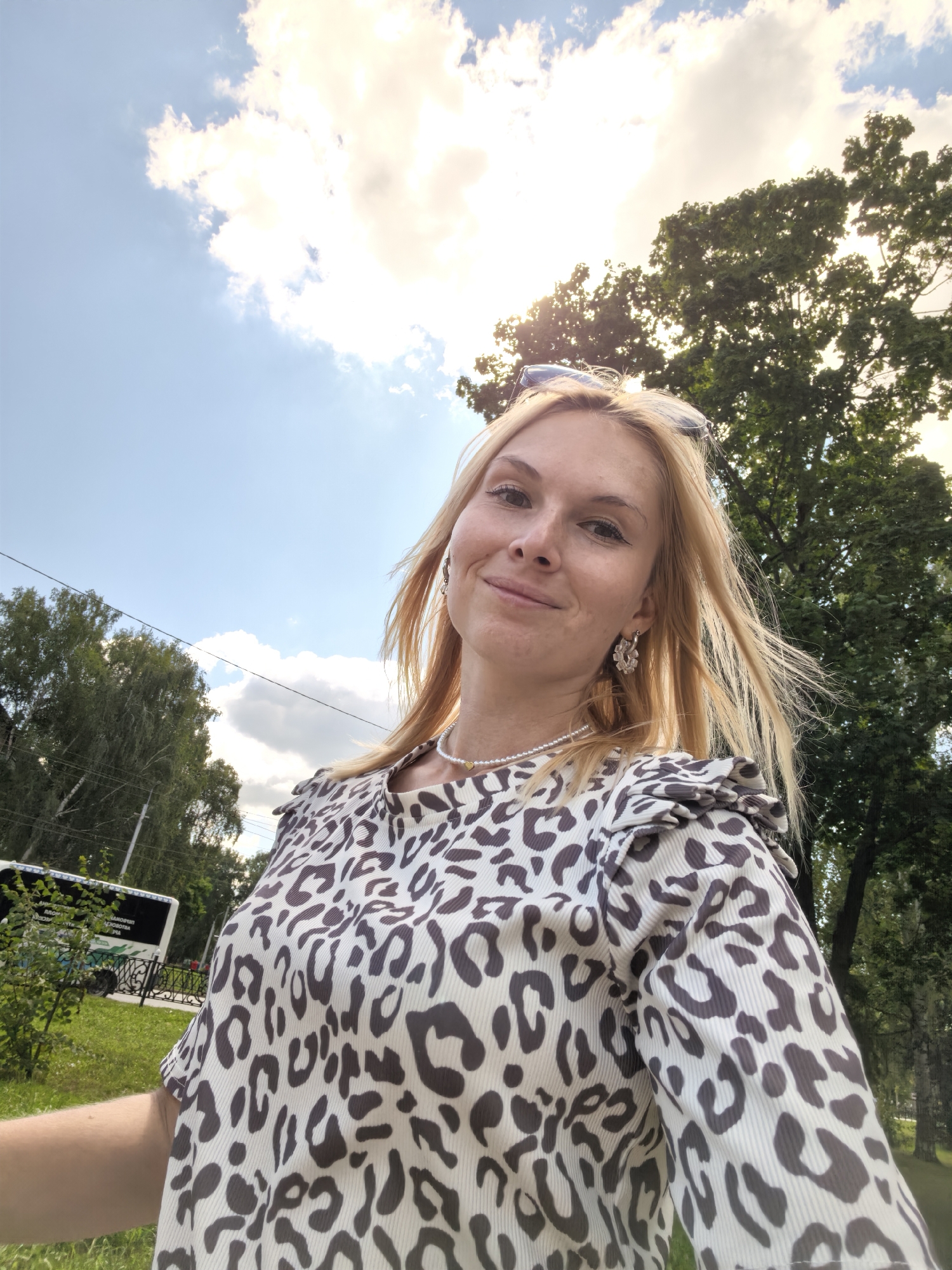 A blonde woman wearing a leopard-print top and a pearl necklace is taking a selfie outdoors. She has blonde hair, blue eyes, and is smiling. Sunglasses are perched on her head. She is standing in a park-like setting with trees and a bright sky with clouds in the background. A bus can be seen on the left side of the image, suggesting an urban environment. The scene appears to be in Sterlitamak, Russia, based on the bus's signage. The lighting suggests it is daytime, likely late afternoon or early evening given the warm light.